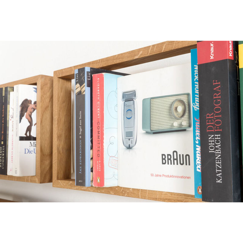 A wooden bookshelf with stainless steel angles showcases colorful books alongside a vintage Braun electric razor box. This vibrant book collection contrasts sharply with the minimalist packaging of the b 8.7 by Das kleine b, blending modern and retro aesthetics.
