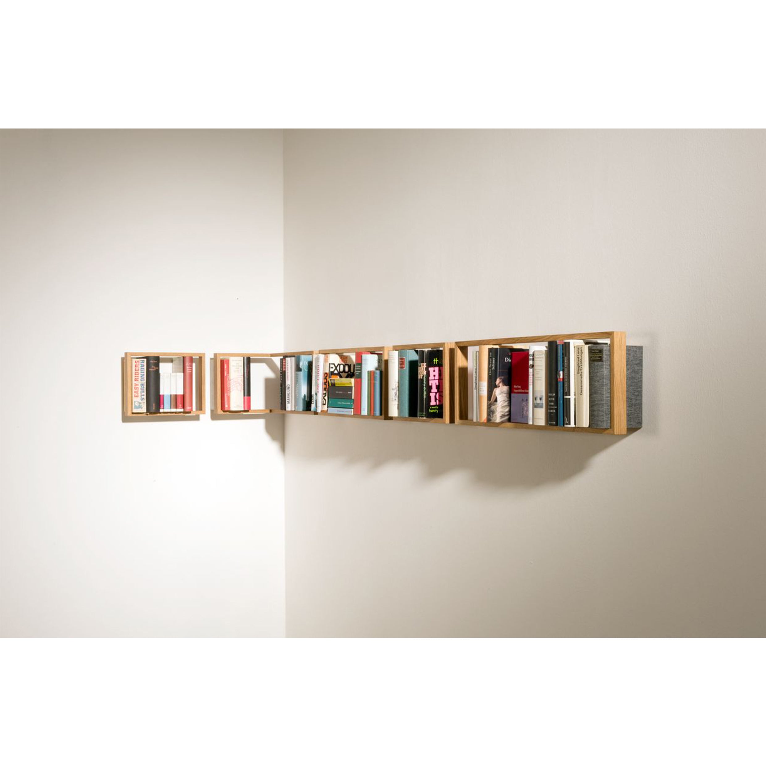 The b corner by Das kleine b features a minimalist design with a wooden frame, offering six square compartments. Perfectly mounted on a pristine white wall, it organizes books into an eye-catching display of colorful spines.
