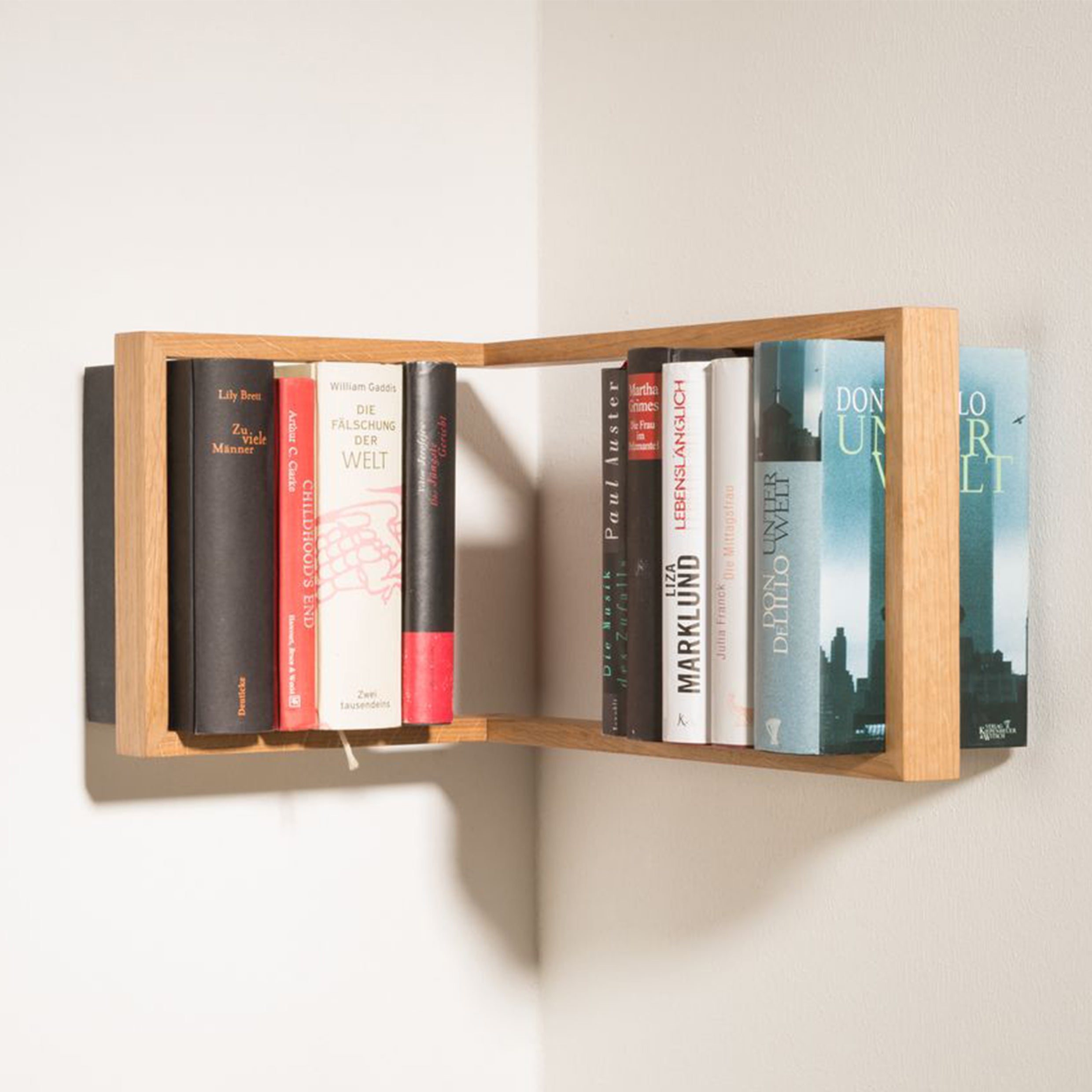 Das kleine bs b corner is a minimalist, L-shaped wooden bookshelf mounted in a corner with a solid frame. Made with light wood and stainless steel angles, it holds books of various titles and colors, creating an intriguing focal point against the wall.