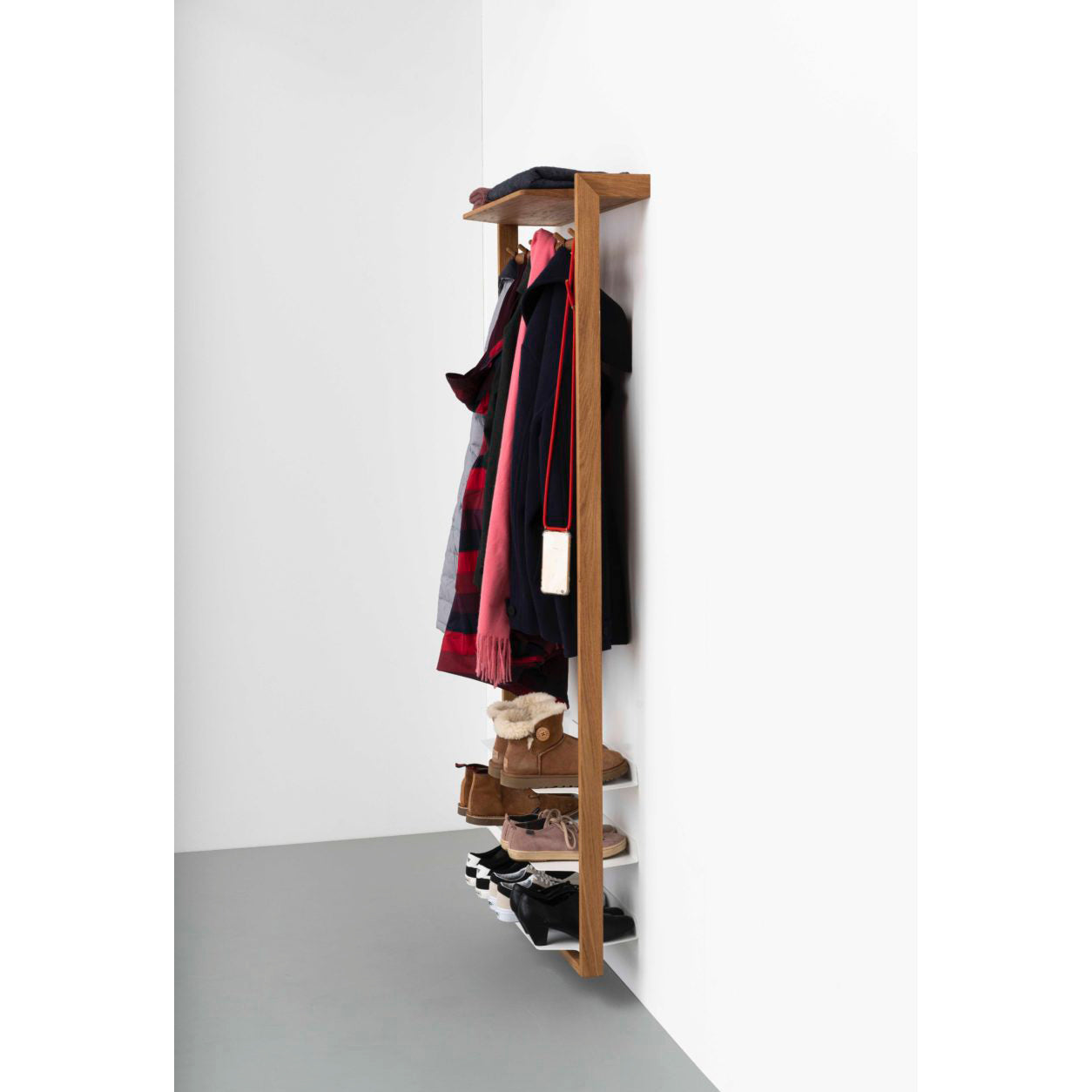 The Gardero-b with Hook Rail by Das kleine b is an oiled oak wall-mounted rack. It stylishly holds a red and black checkered jacket, a black coat, a bag, brown and black shoes on shelves below, and features a cap on the top shelf. The floor is gray with a crisp white wall backdrop.