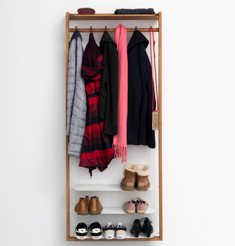 The Das kleine b Gardero-b with Hook Rail is an elegant corridor piece crafted from oiled oak, featuring hooks adorned with coats and scarves. Shelves below hold sneakers, boots, and heels. A hat and small bag rest on top as it stands against a plain white wall.