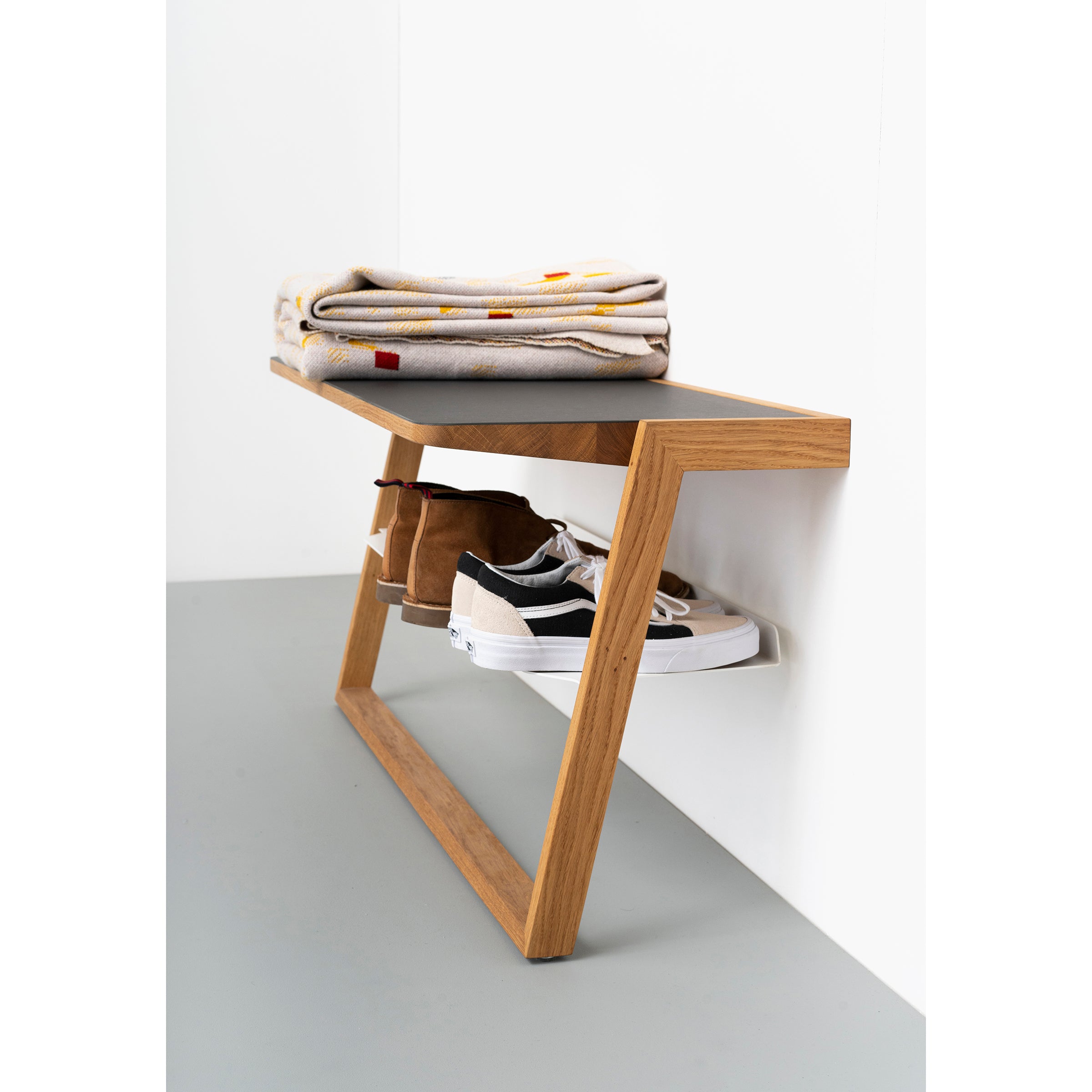 The Bench Bänkle by Das kleine b, a minimalist wooden shoe rack, leans against a white wall topped with folded blankets. Brown boots and white sneakers are neatly arranged below it. The gray floor enhances its sleek, modern design, making it perfect as space-saving furniture.