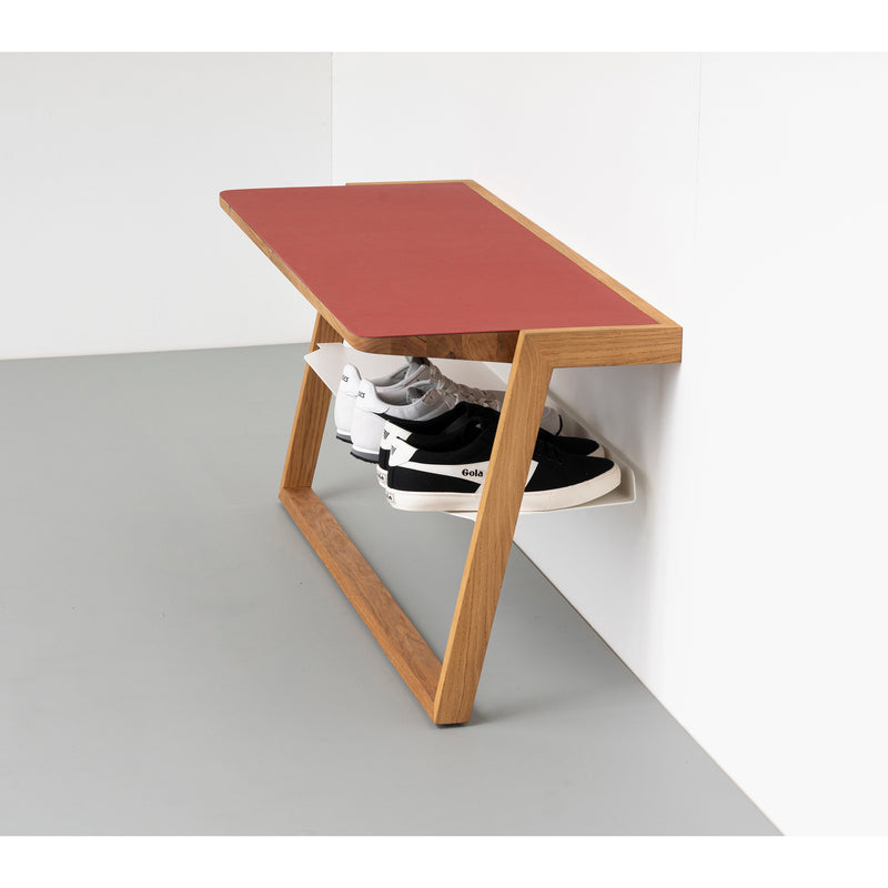 The Bench Bänkle by Das kleine b is a minimalist wall-mounted shoe rack with a red top, featuring a zigzag design for space-saving. It effortlessly holds black and white sneakers underneath, complemented by a sleek light gray floor.