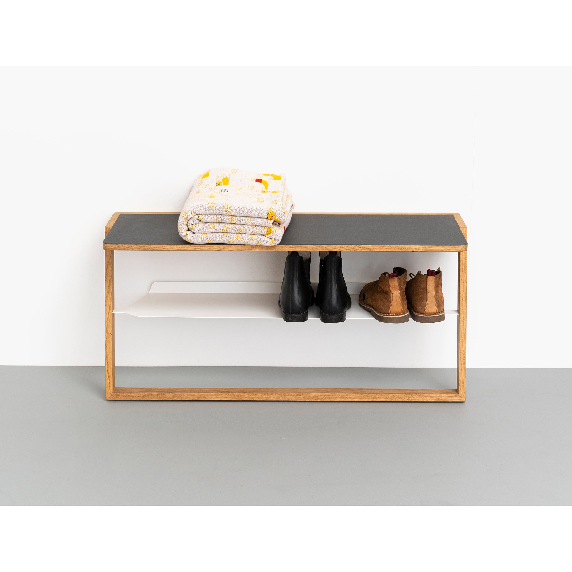 The Bench Bänkle by Das kleine b features a wooden frame with a black cushion, positioned against a white wall. It hosts a folded blanket, while its aluminum shelf holds three pairs of shoes, including black and brown boots—ideal space-saving furniture for any entryway.