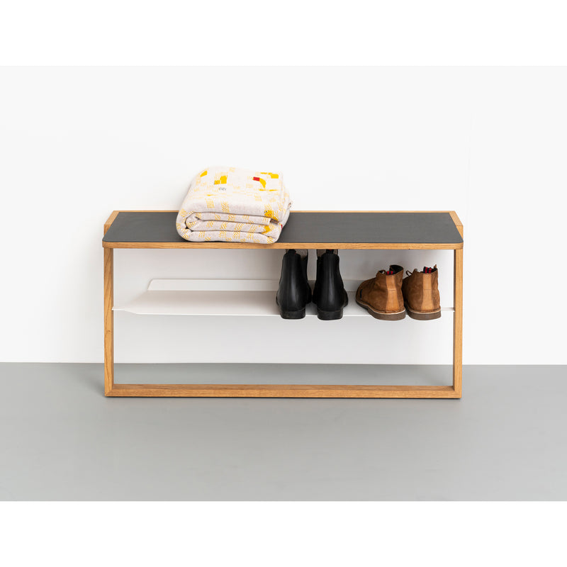 The Bench Bänkle by Das kleine b features a wooden frame with a black cushion, positioned against a white wall. It hosts a folded blanket, while its aluminum shelf holds three pairs of shoes, including black and brown boots—ideal space-saving furniture for any entryway.