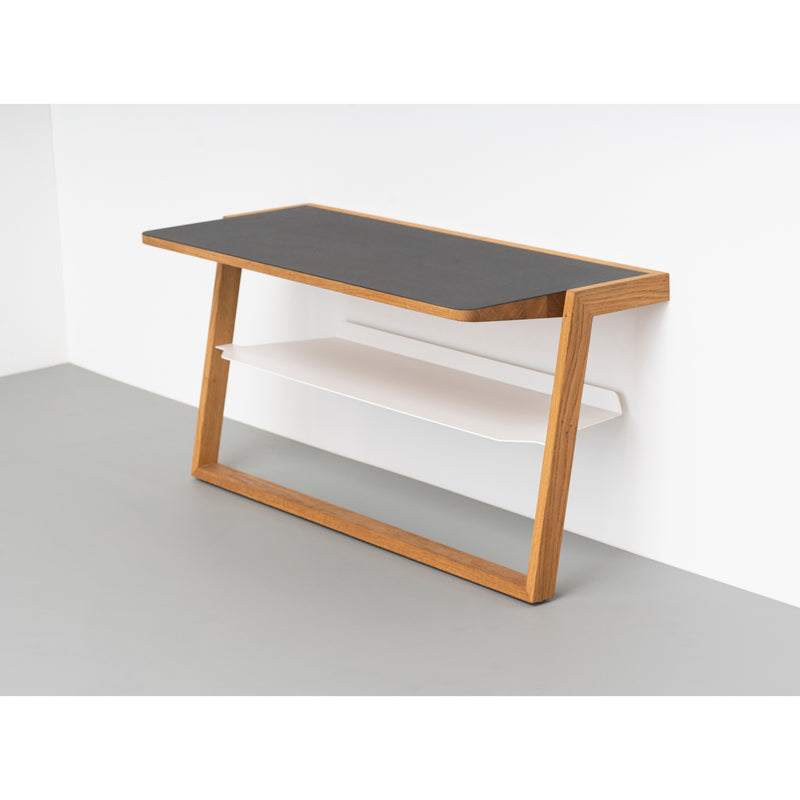 The Bench Bänkle by Das kleine b is a minimalist wooden desk with a black surface and angular frame, featuring a space-saving white shelf underneath. Set against a white wall on a gray floor, it embodies efficient space usage.