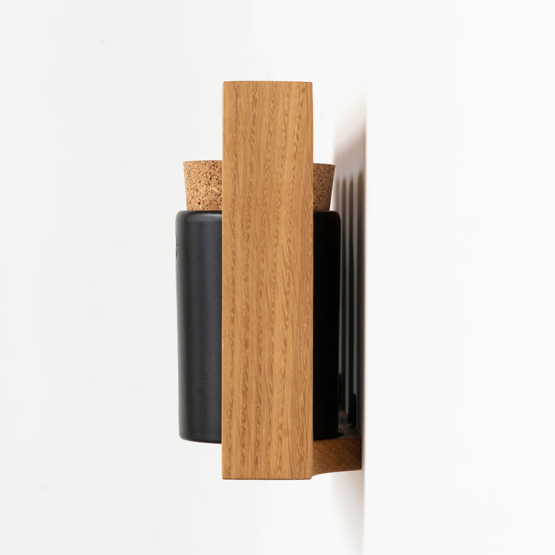 Side view of the Spice Rack Bocuse by Das kleine b, a modern, wall-mounted shelf crafted from light oak. It features a minimalist design with a black cylindrical container and cork lid, set against a plain white background.
