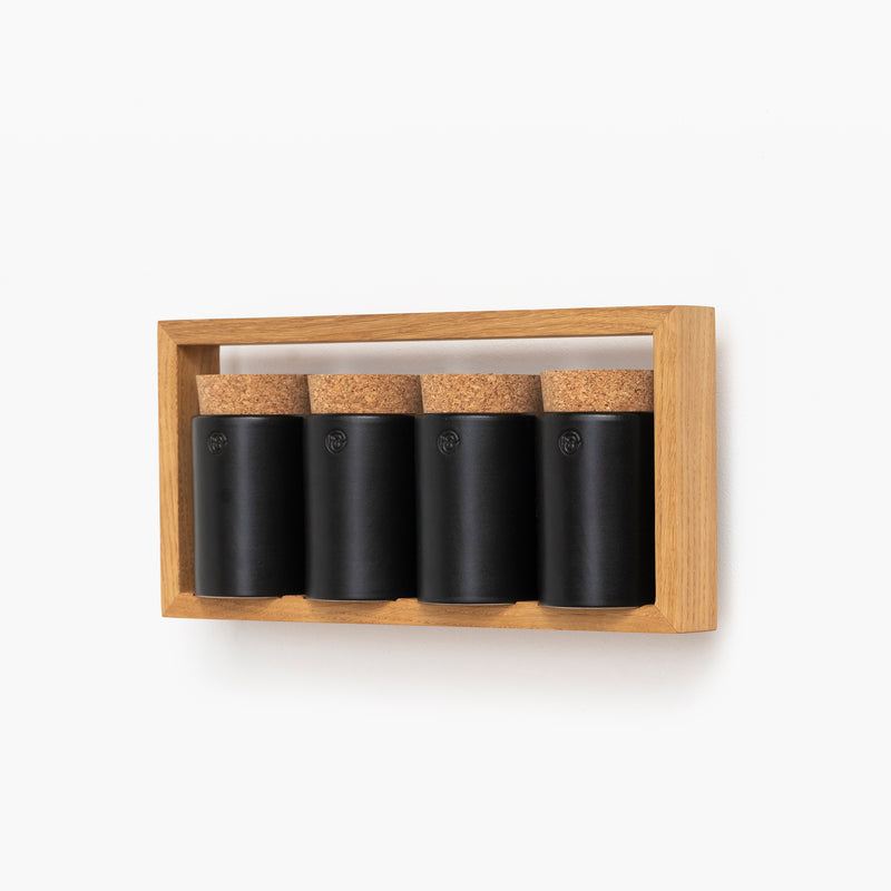Four black cylindrical Spice Rack Bocuse jars with cork lids from Das kleine b are neatly arranged in a horizontal oak frame against a white background.