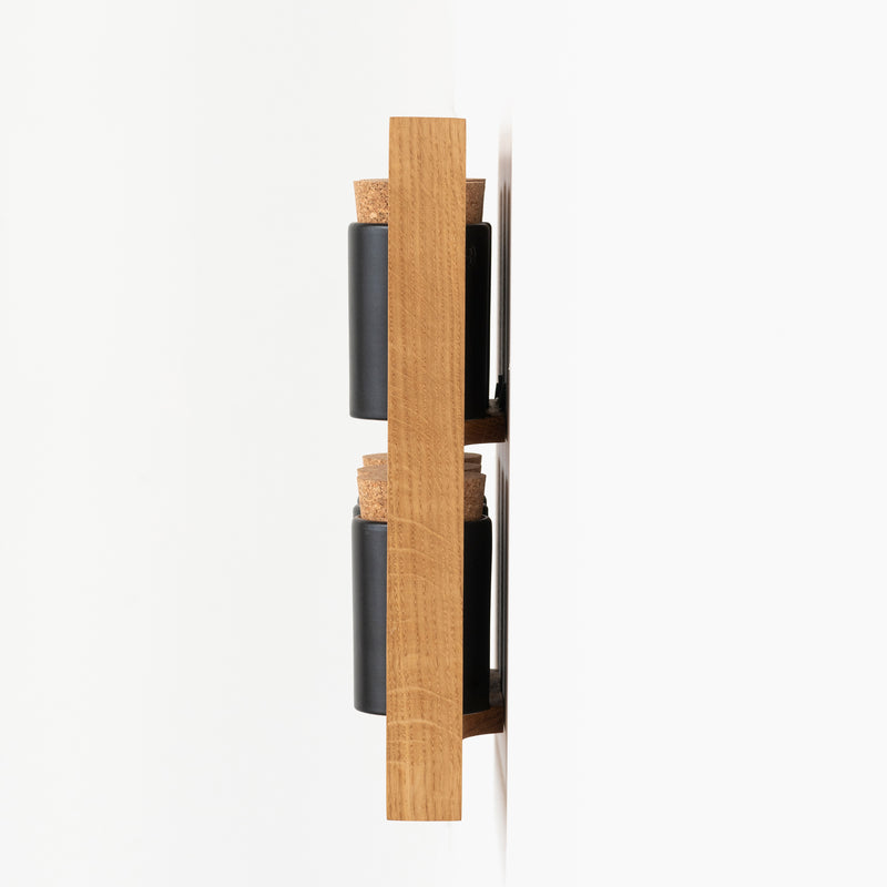 Side view of two black and cork Spice Rack Bocuse jars by Das kleine b, held in a vertical oak bracket mounted on a white wall. The modern, minimalist design showcases the aligned and partially visible spice racks.