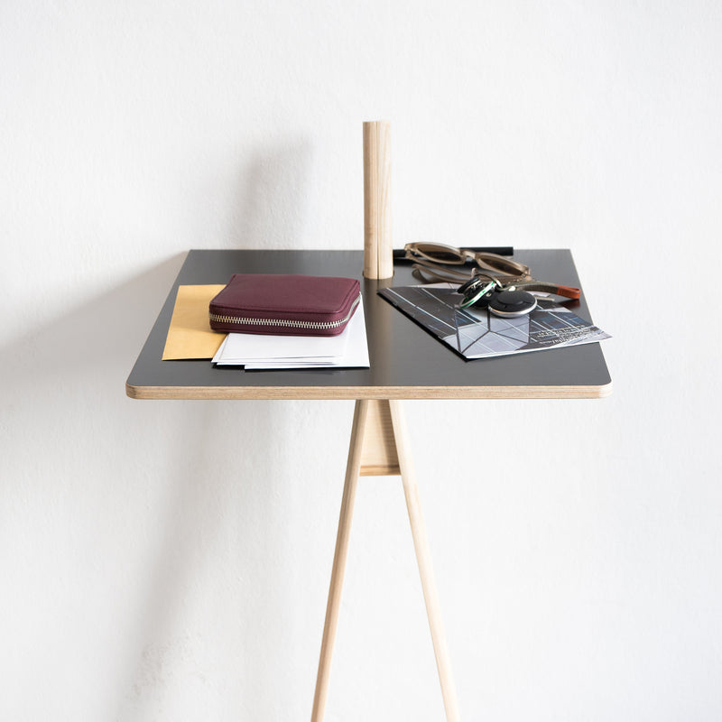 The Moormann Der Kleine Lehner writing stand, with a minimalist black surface, elegantly showcases sunglasses, a wooden block, papers, a brown wallet, and an envelope against a white wall.