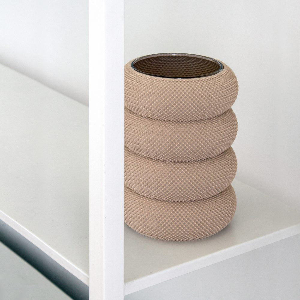 The Dorn 341g 1c Vase by Sheyn, eco-friendly and cylindrical with a stacked ring design, sits on a white shelf. Crafted through 3D printing, it features an intricate pattern and offers a modern minimalist look against the stark background.