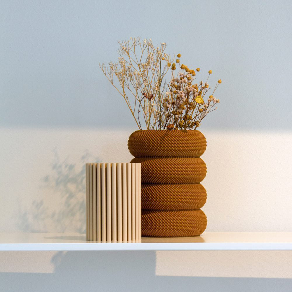 A Dorn 341g 1c Vase by Sheyn, textured in brown and filled with dried flowers, pairs elegantly with a ribbed beige vase on a white shelf against a pale blue wall. The setting crafts an eco-friendly ambiance as soft shadows cast left, showcasing the artistry of a design studio.