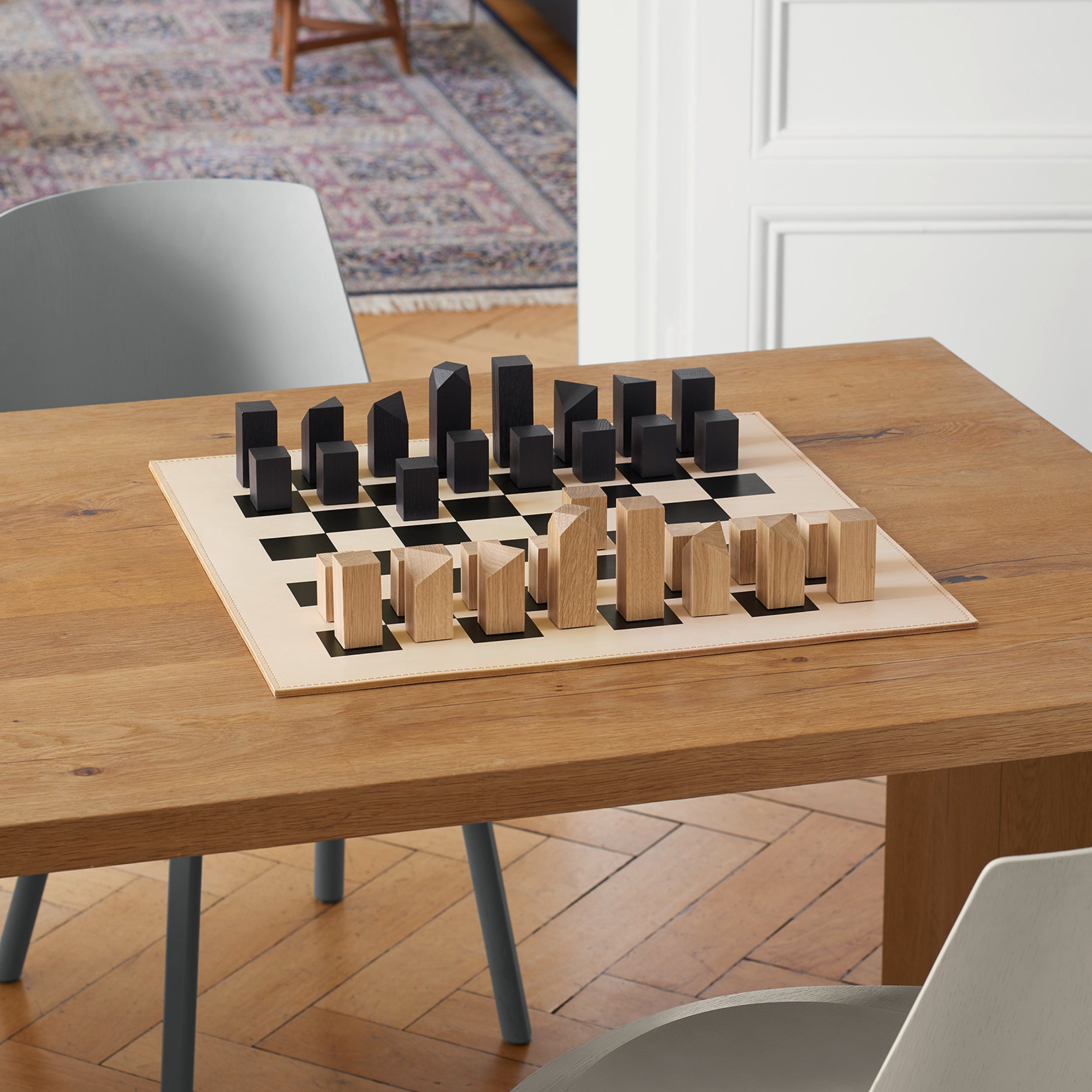 The AC03 NONA chess set by E15, featuring geometric wooden figures on a leather board, adorns a wooden dining table surrounded by light gray chairs. Handmade in Germany, it complements the rooms charm alongside a stylish rug and elegant wooden floor.
