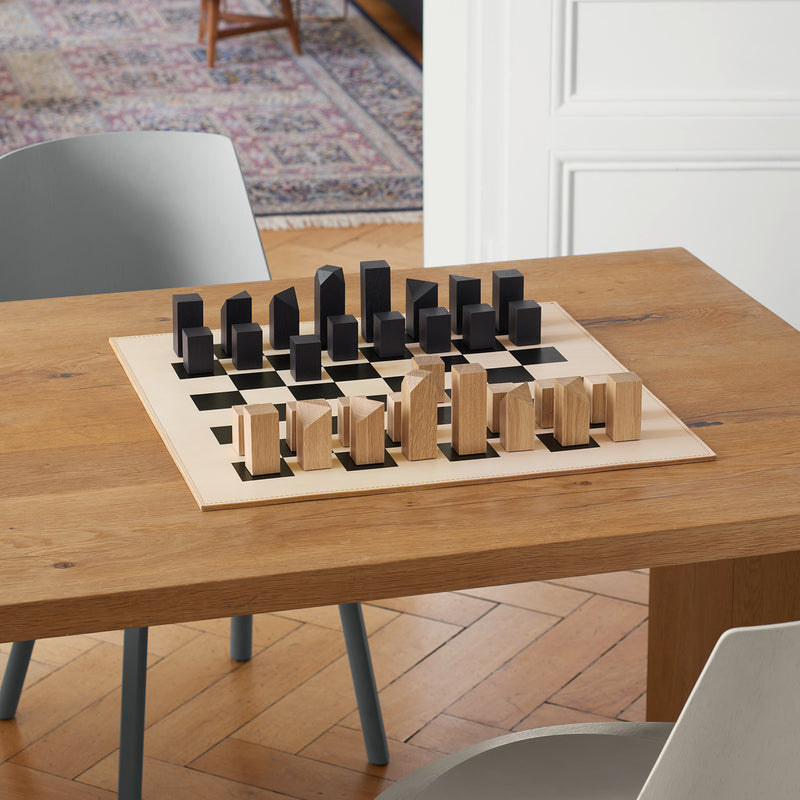 The AC03 NONA chess set by E15, featuring geometric wooden figures on a leather board, adorns a wooden dining table surrounded by light gray chairs. Handmade in Germany, it complements the rooms charm alongside a stylish rug and elegant wooden floor.