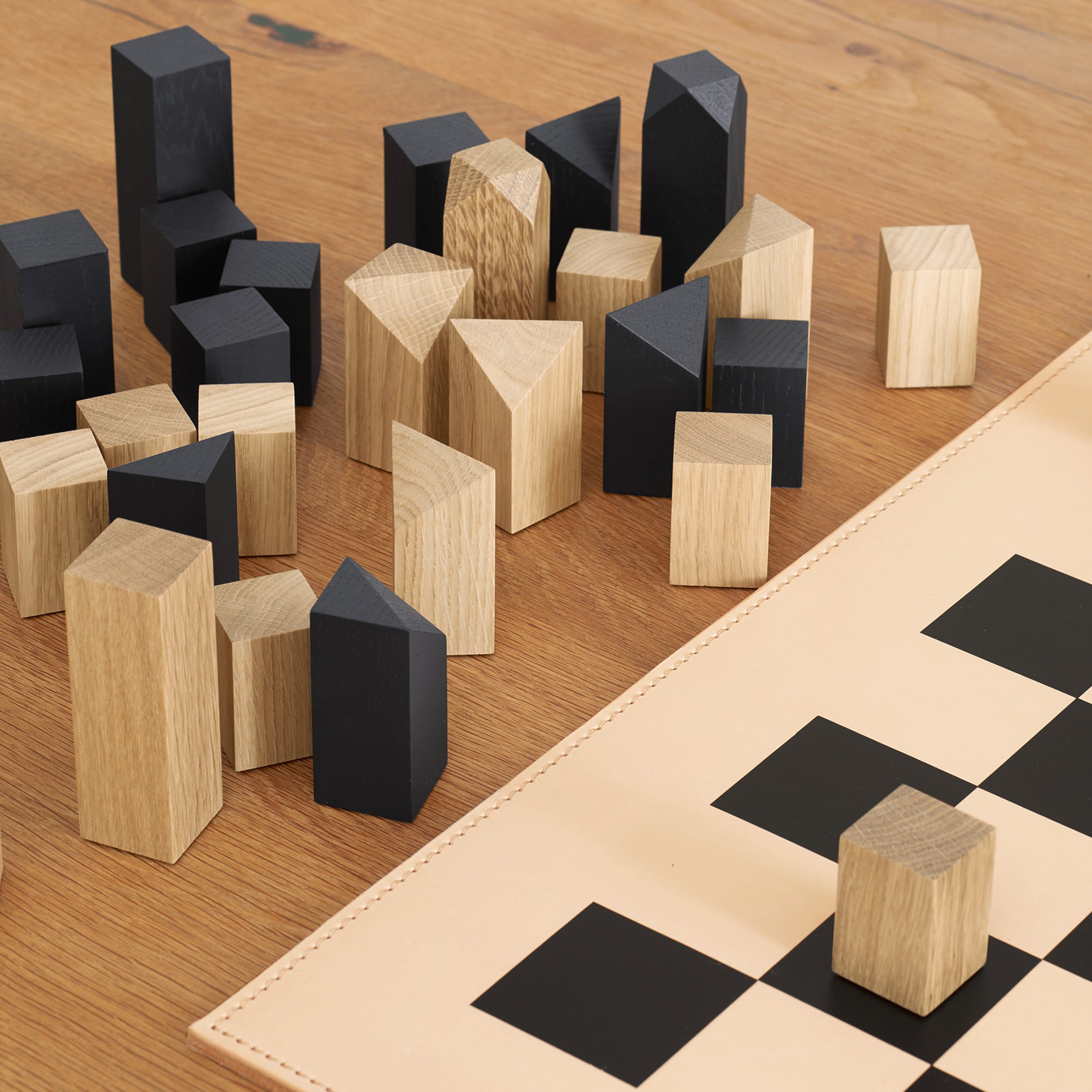 Wooden cubes and triangular prisms in natural and black finishes are scattered on a wooden table beside an AC03 NONA CHESSBOARD AND FIGURES from E15. One small, handmade German wooden cube is placed on the boards corner.
