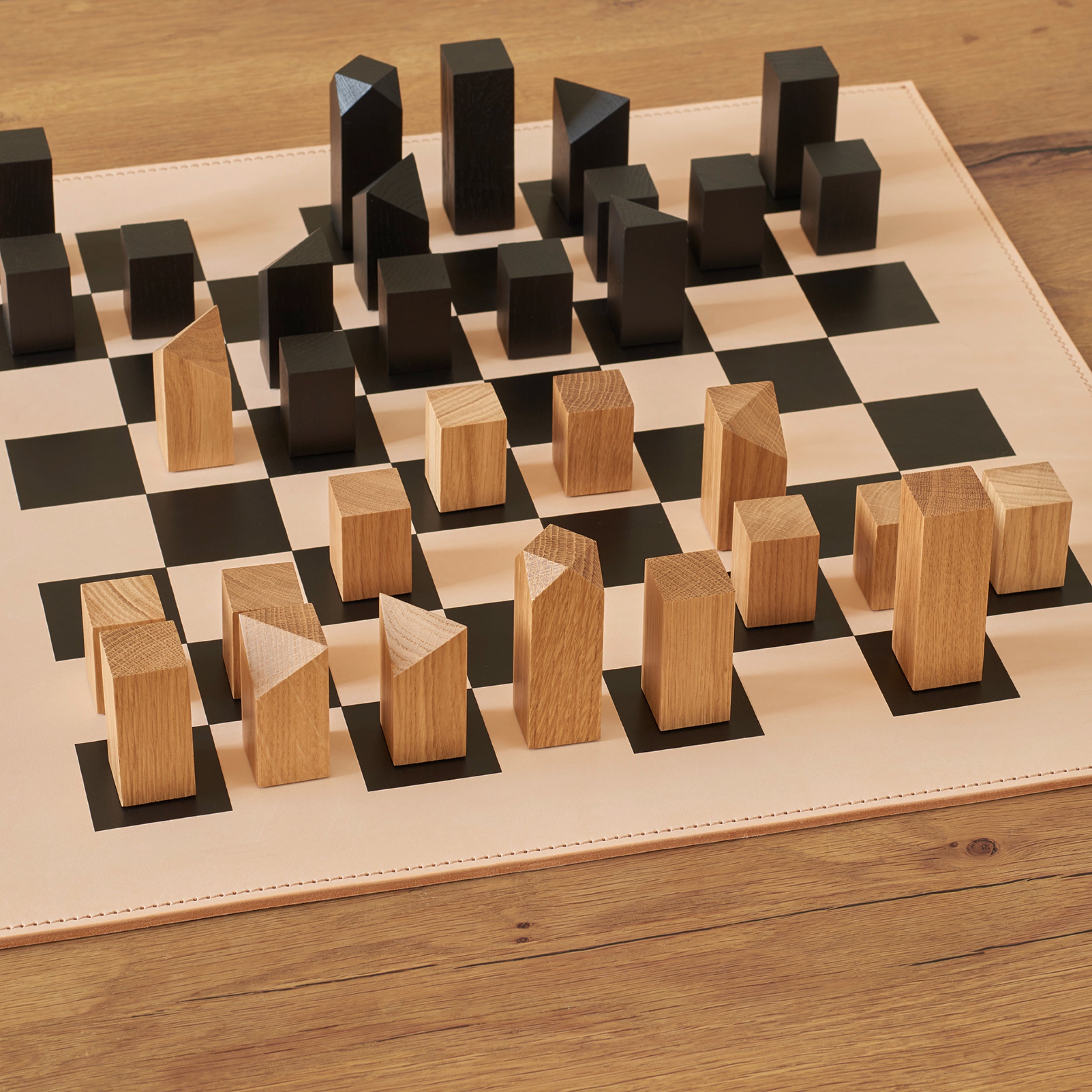 The AC03 NONA Chessboard and Figures by E15 features a modern design with minimalist, rectangular wooden pieces in light and oak tones, arranged on a black and cream grid on a wooden table surface.