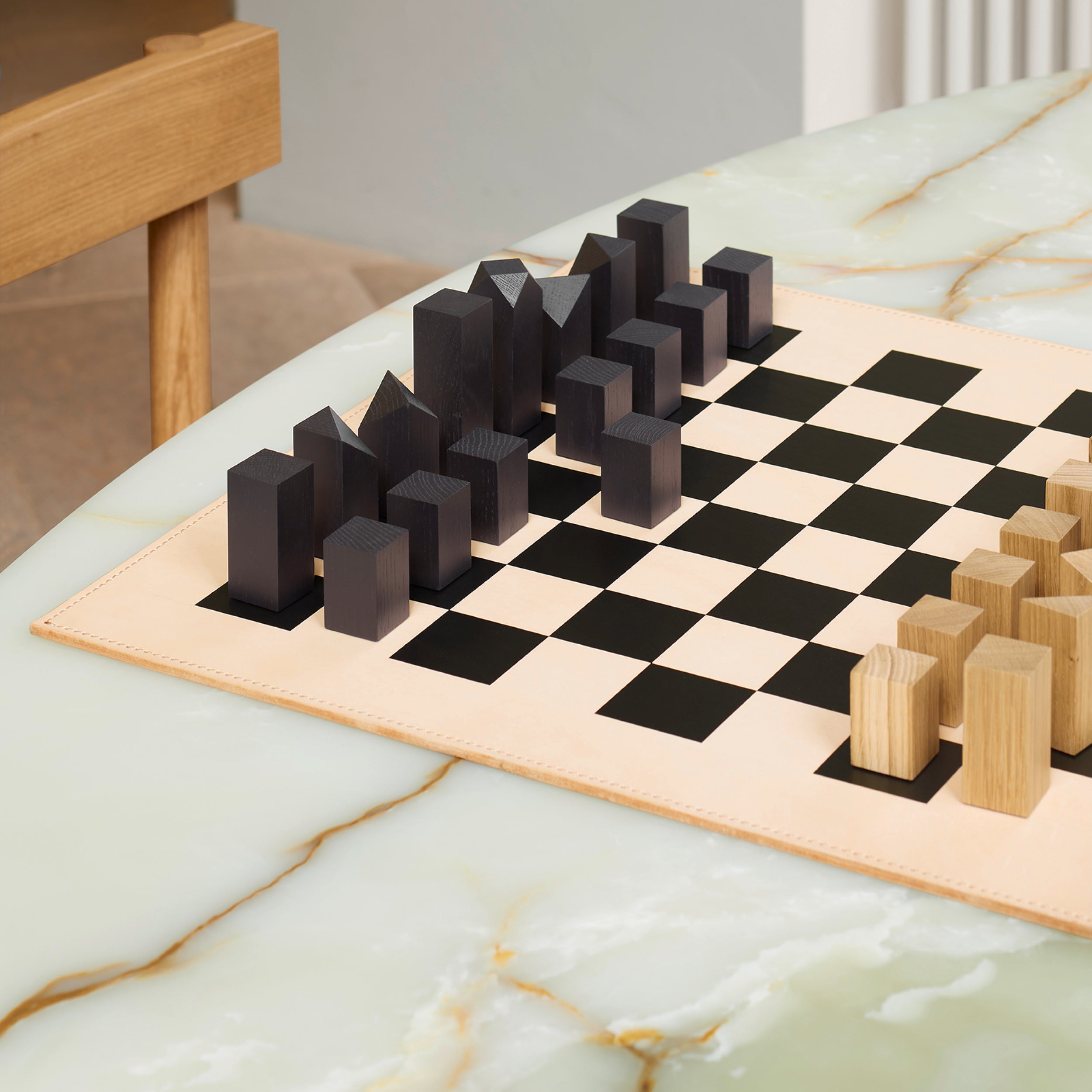 The AC03 Nona Chessboard and Figures by E15 features geometric chess pieces elegantly displayed on a marble table, with black ones on the left and wooden ones on the right, perfectly complementing a partially visible wooden chair. Handmade in Germany.