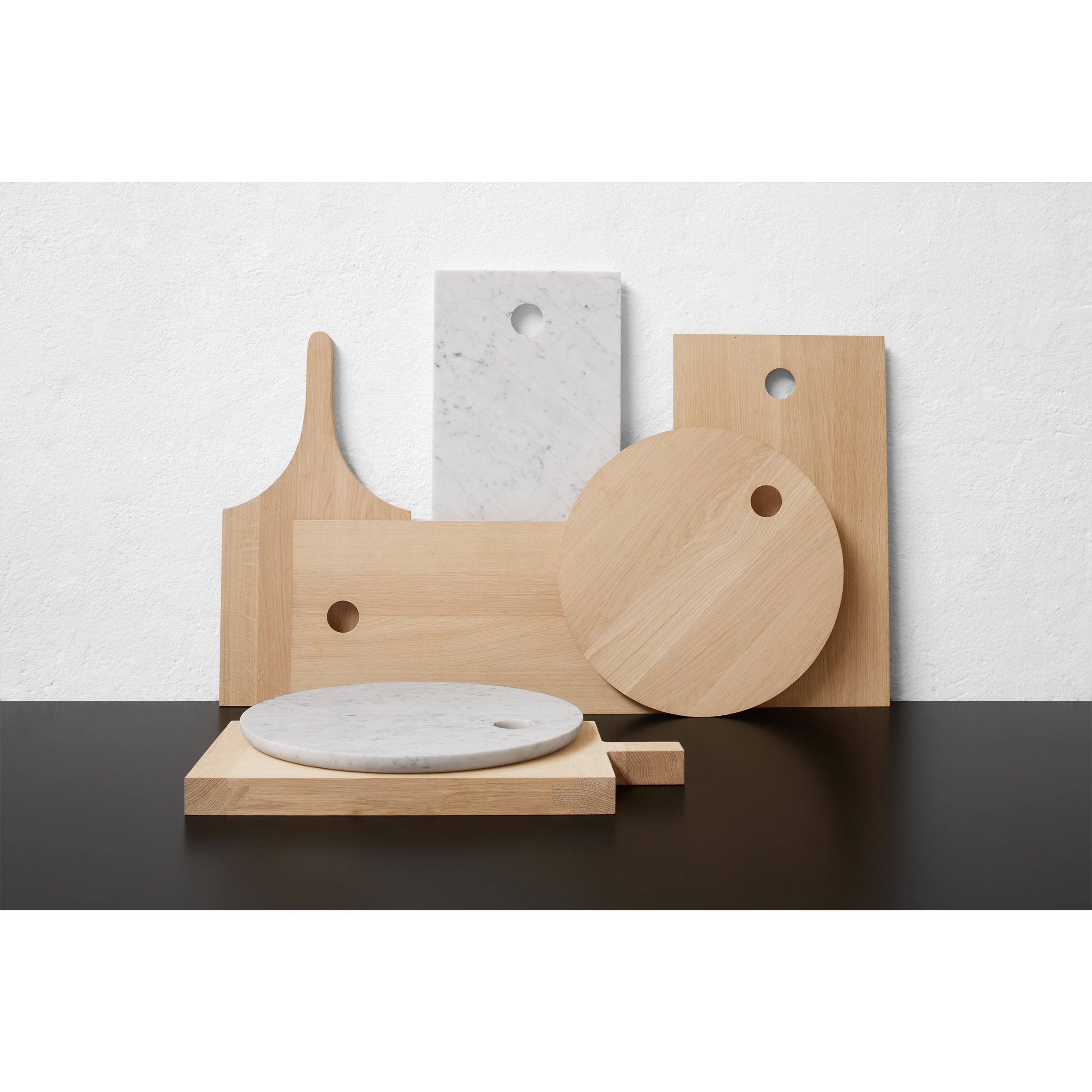 E15s AC07 CUT collection features untreated oak and marble cutting boards in circular, rectangular, and paddle shapes, displayed upright against a white textured wall on a dark surface.