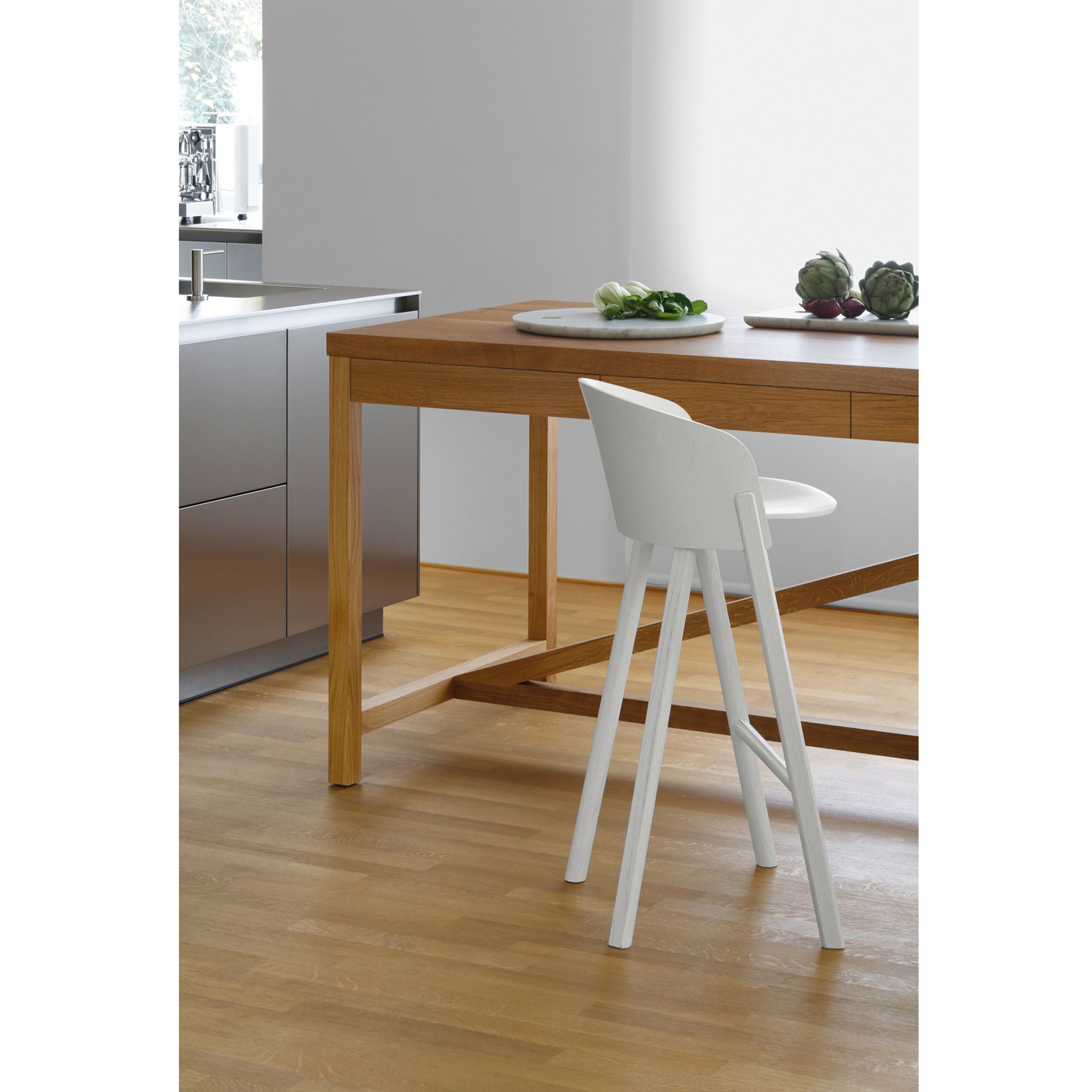A minimalist kitchen scene showcases an E15 AC07 CUT wooden dining table made from untreated oak, paired with a sleek white chair. Artichokes and greens sit atop the table on a solid wood floor, while a stainless steel kitchen counter with sink and faucet is seen in the background.
