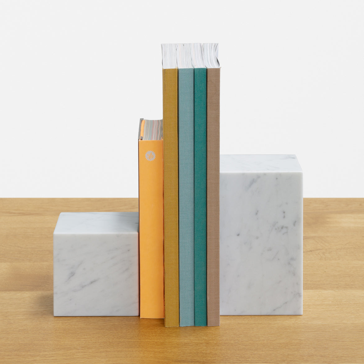 Three books with vibrant orange, teal, and tan covers stand against E15 AC11 STOP white marble blocks of varying heights on a wooden surface, creating a modern, minimalist contrast.