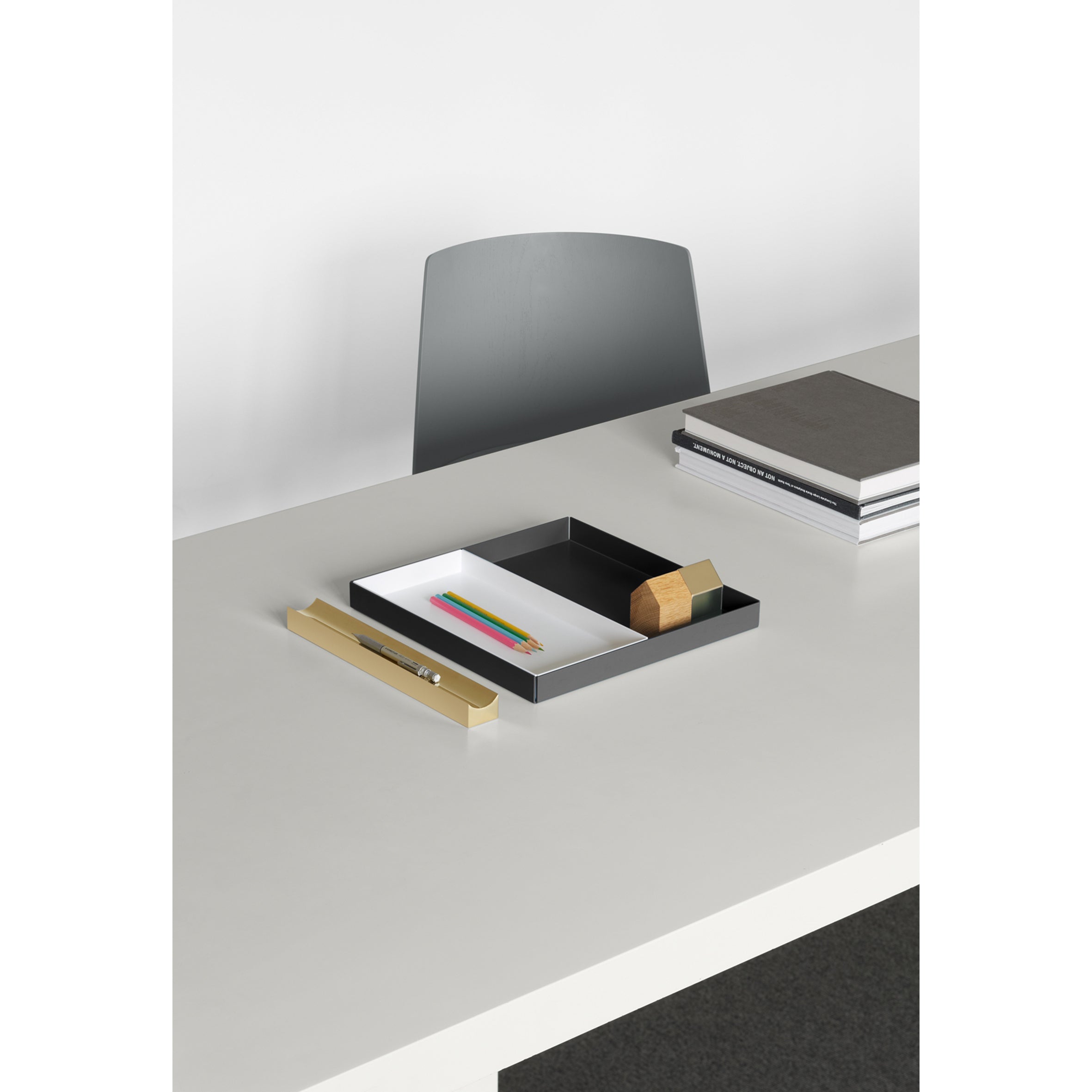 A minimalist desk with a gray chair features a tray with colored pencils, wooden blocks, and stationery. Nearby, neatly stacked books and an E15 AC12 PEN rest against the white wall backdrop.