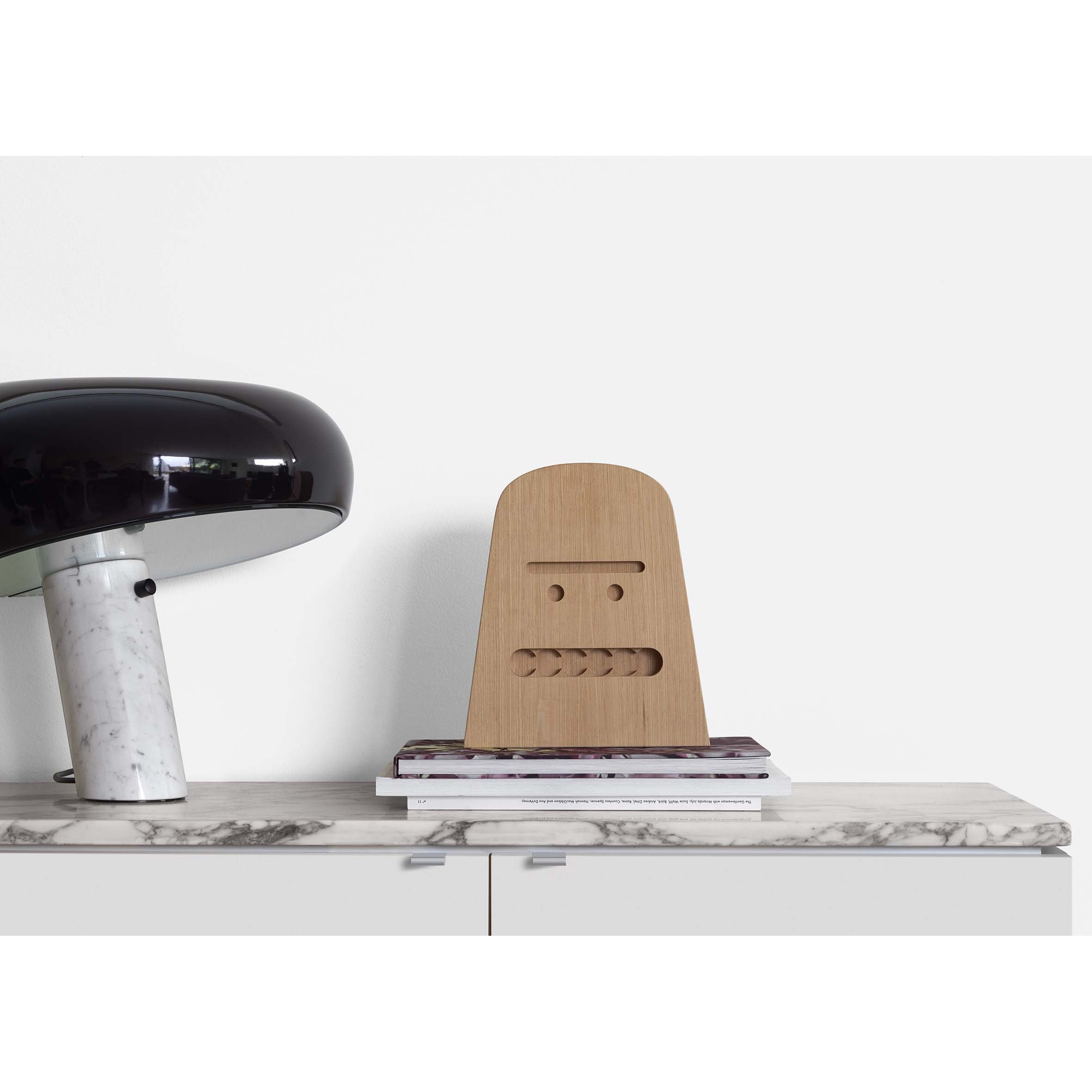 An E15 AC17 BIG B lamp with a black dome shade and marble base rests on an untreated oak-top cabinet. Beside it is a solid wood robot face sculpture atop stacked magazines against a plain white wall.