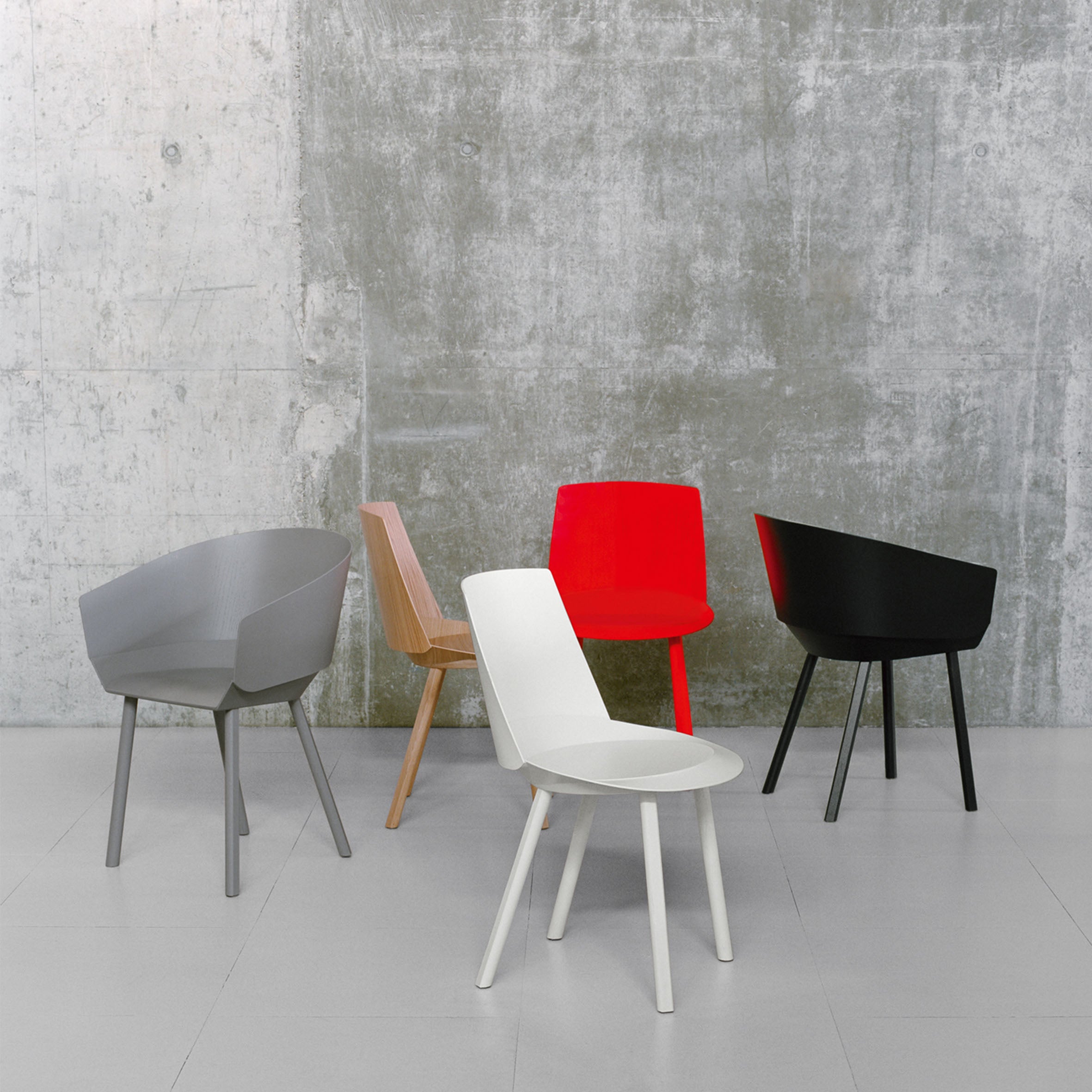 A set of E15s CH04 HOUDINI Armchairs in a minimalist design, with lacquered plywood in gray, wood, red, white, and black hues, is arranged on a light gray floor against a textured concrete wall.