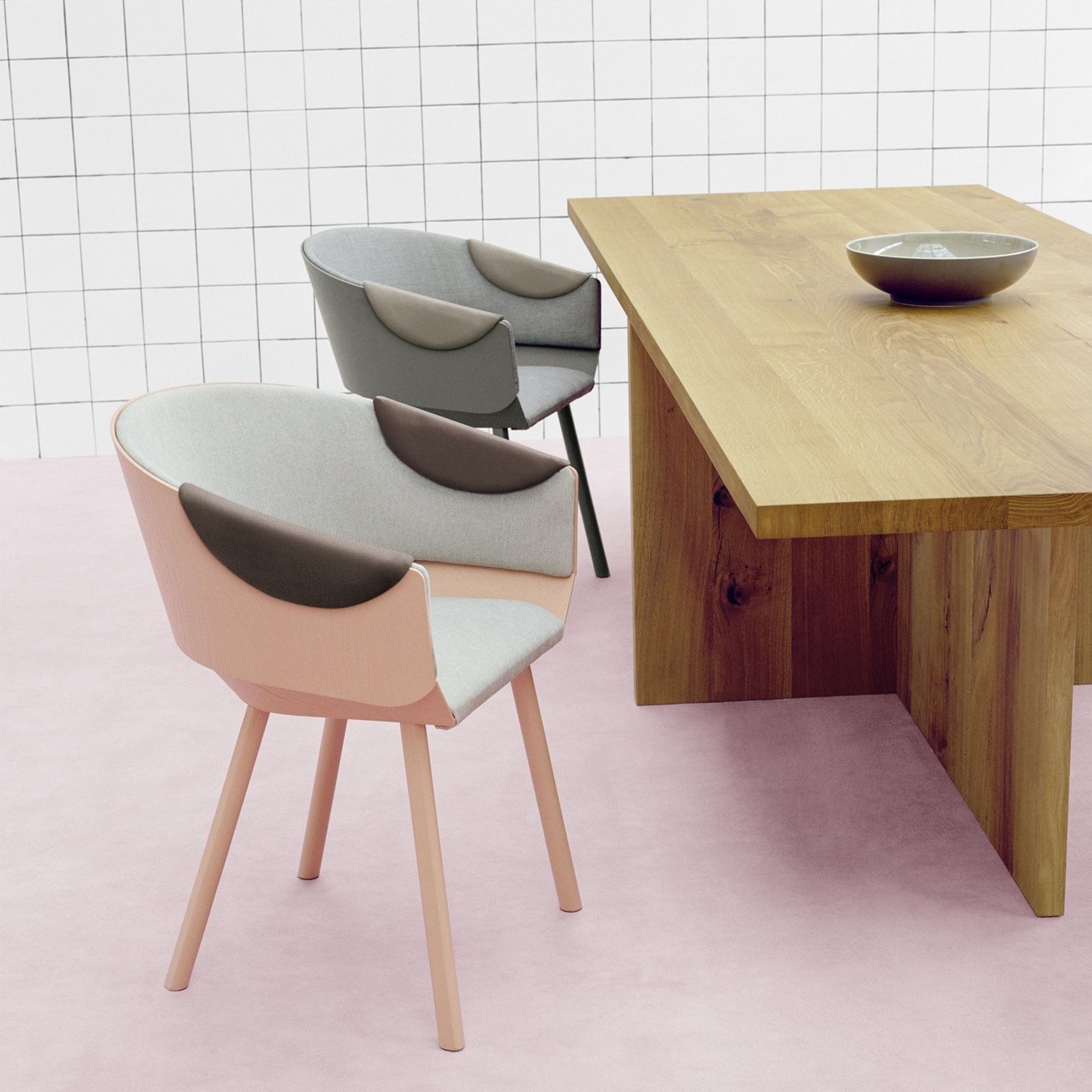 A modern dining area features two CH04 HOUDINI Armchairs by Stefan Diez for E15, with soft gray and muted pink upholstery. They surround a light wood rectangular table topped with a decorative bowl, set against a white tiled wall and a pale pink lacquered plywood floor.