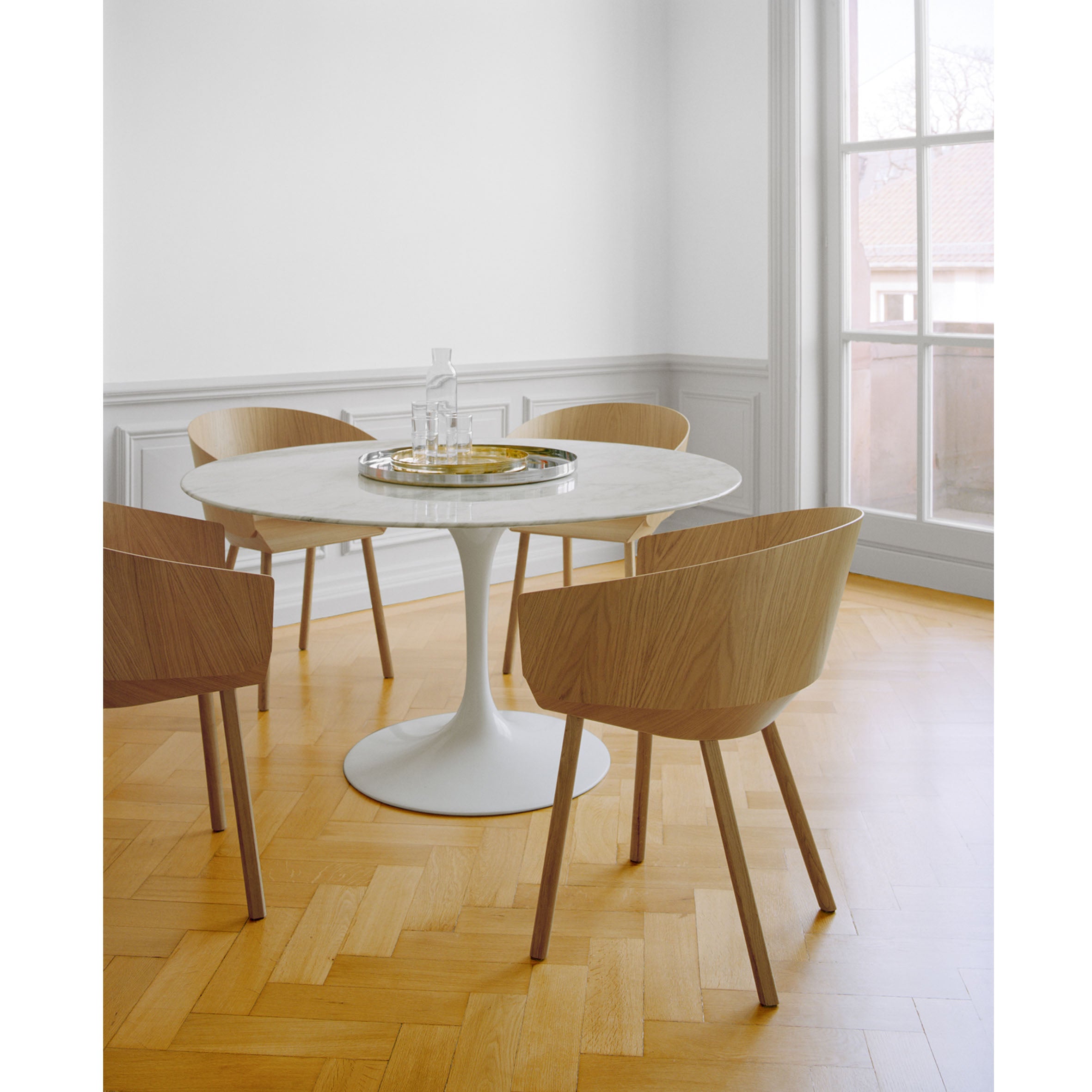 A minimalist dining room showcases a round white table with four E15 CH04 HOUDINI Armchairs designed by Stefan Diez, made from lacquered plywood. A glass carafe and glasses rest on a tray. Light wood herringbone flooring and natural light enhance the serene ambiance.