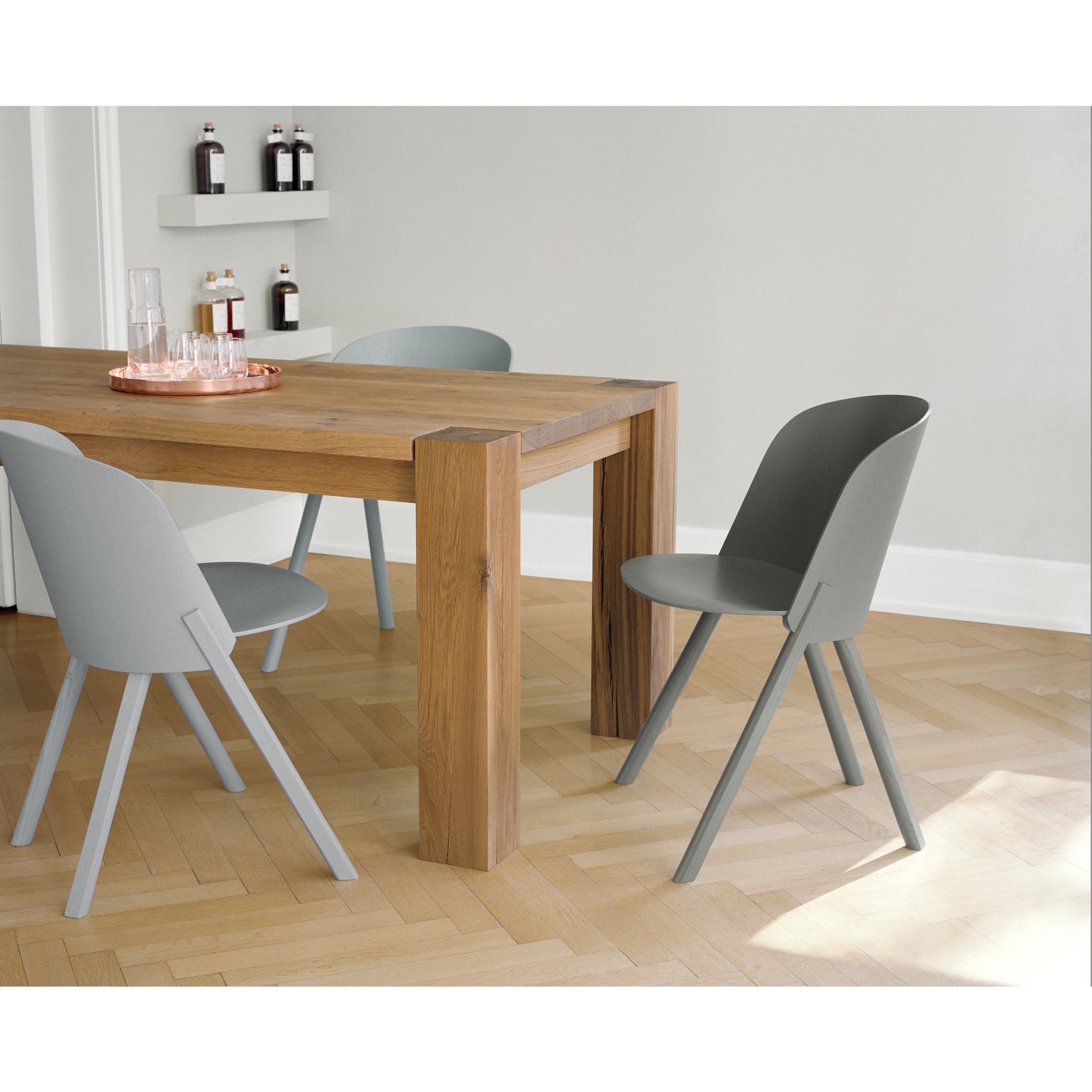 A modern dining area highlights an oak veneer plywood table paired with three E15 CH05 THIS Side Chairs in minimalist gray, situated on a light wood parquet floor. Bottles line the white wall shelves, and a tray with glassware adds elegance to the table.