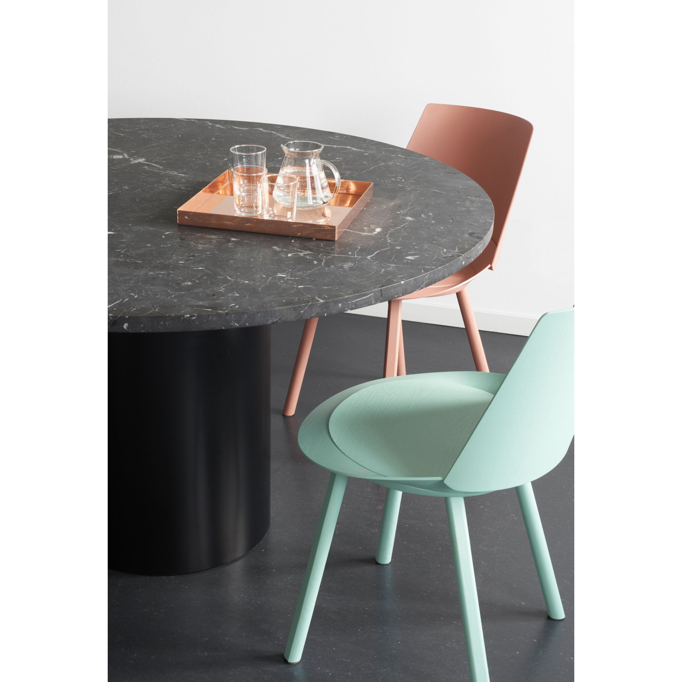 The CM04 ITO by E15 is a round black marble table with a cylindrical base, adorned with a wooden tray holding a glass jug and two glasses. Two powder-coated aluminum chairs, one peach and the other mint green, surround the table on a dark floor.