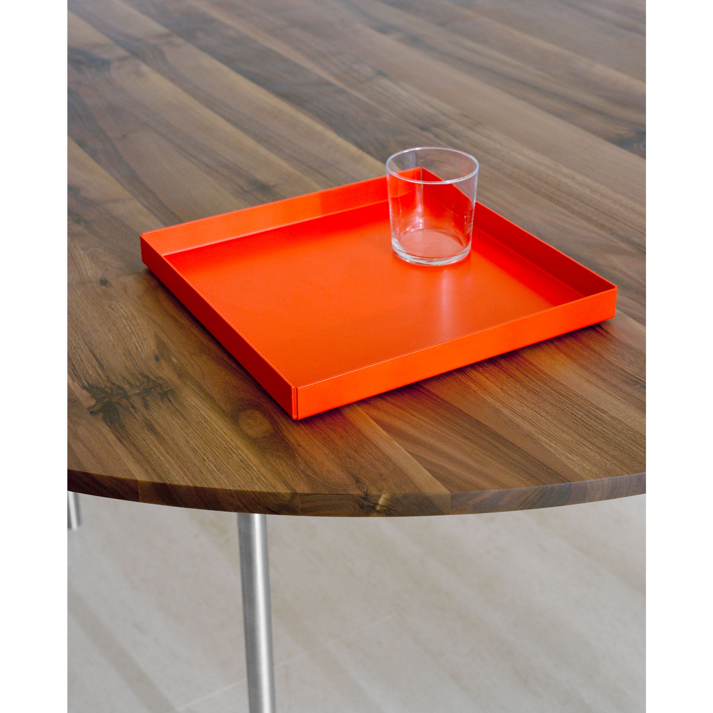 The E15 CM04 ITO clear glass sits on a vibrant orange tray on a round wooden table with a polished surface and stainless steel legs, set against a light-colored floor.
