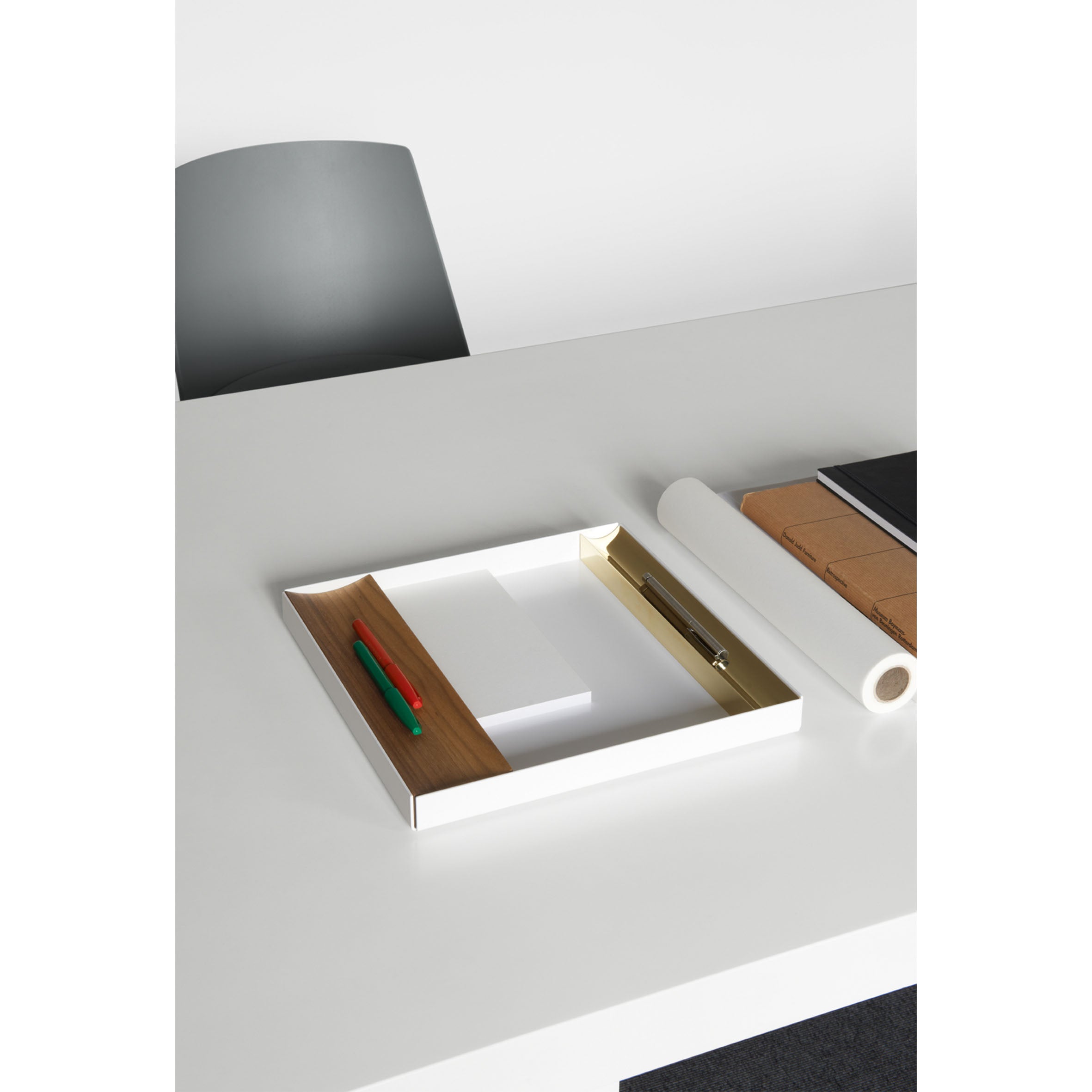 A minimalistic workspace showcases an E15 CM04 ITO white table paired with a beige chair. The table holds a sleek powder-coated aluminum tray, containing red and green pens, a notebook, white paper roll, and a metal ring folder.