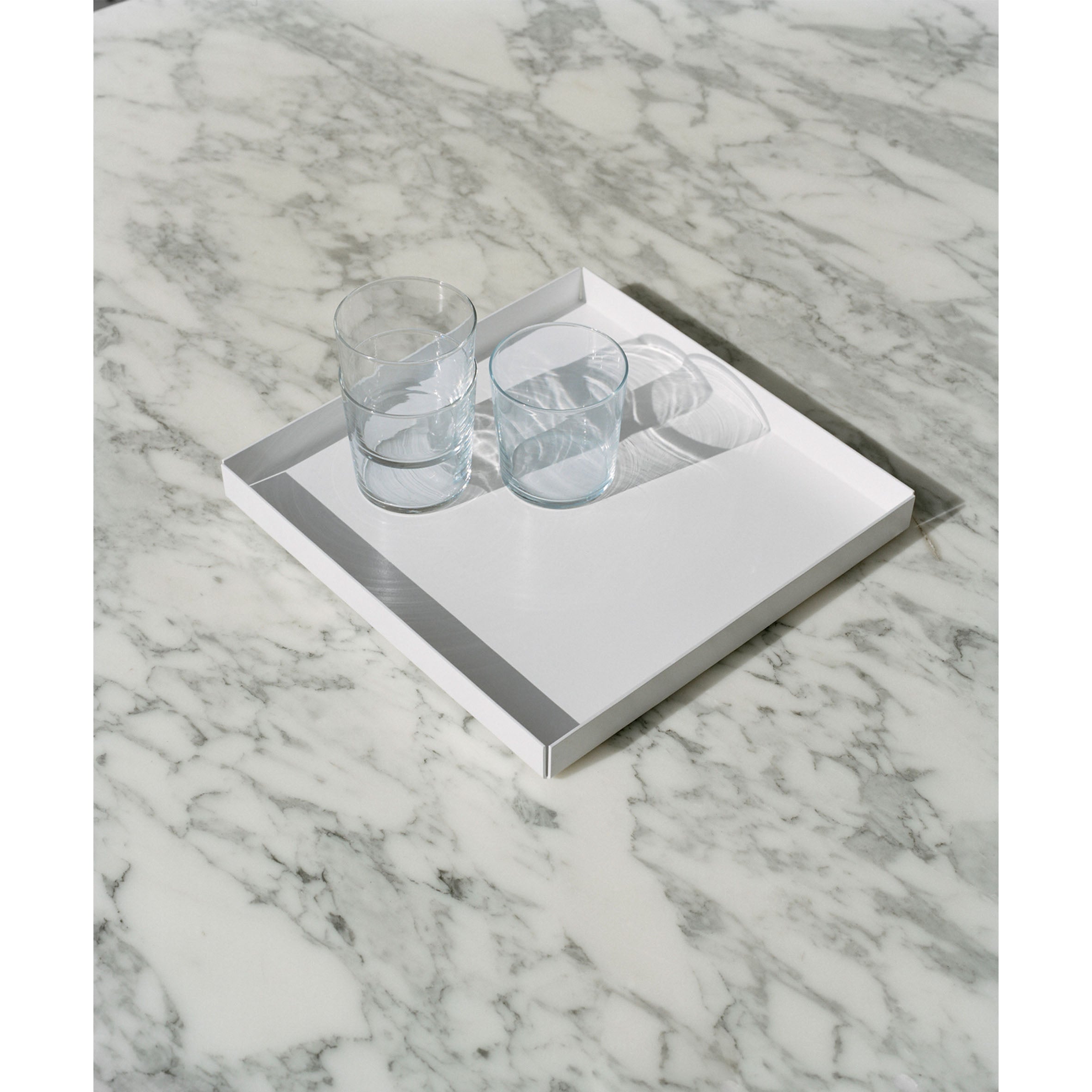 The CM04 ITO by E15 features a white powder-coated aluminum tray with three clear glass cups, set on a light-colored marble surface. The minimal design is orderly as the tray elegantly casts a shadow on the marble.