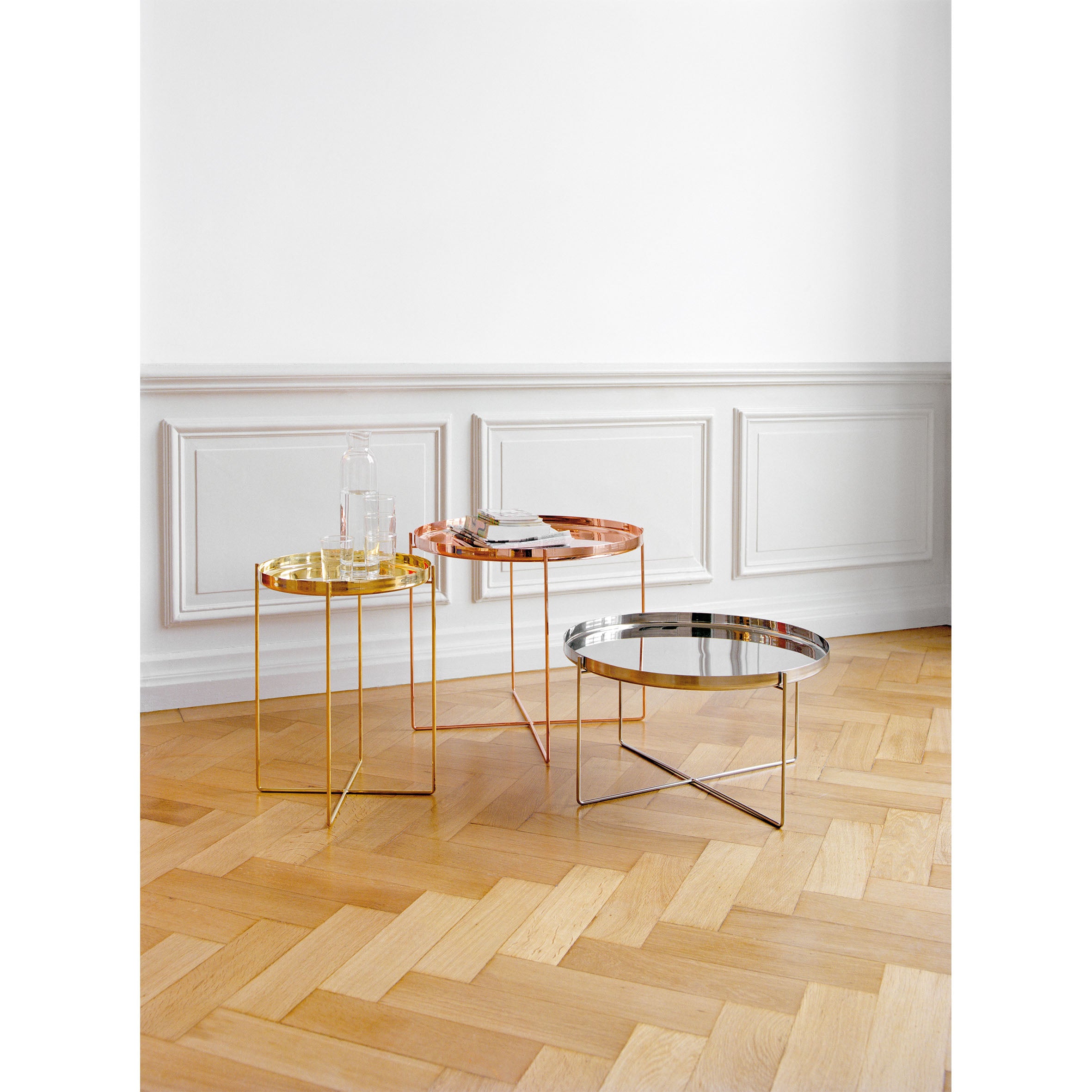 Three modern round CM05 HABIBI TRAY tables by E15, in gold, copper, and silver, stand elegantly on a herringbone wood floor. Each glass-topped table holds an ornamental tray or books against classic white paneled walls.