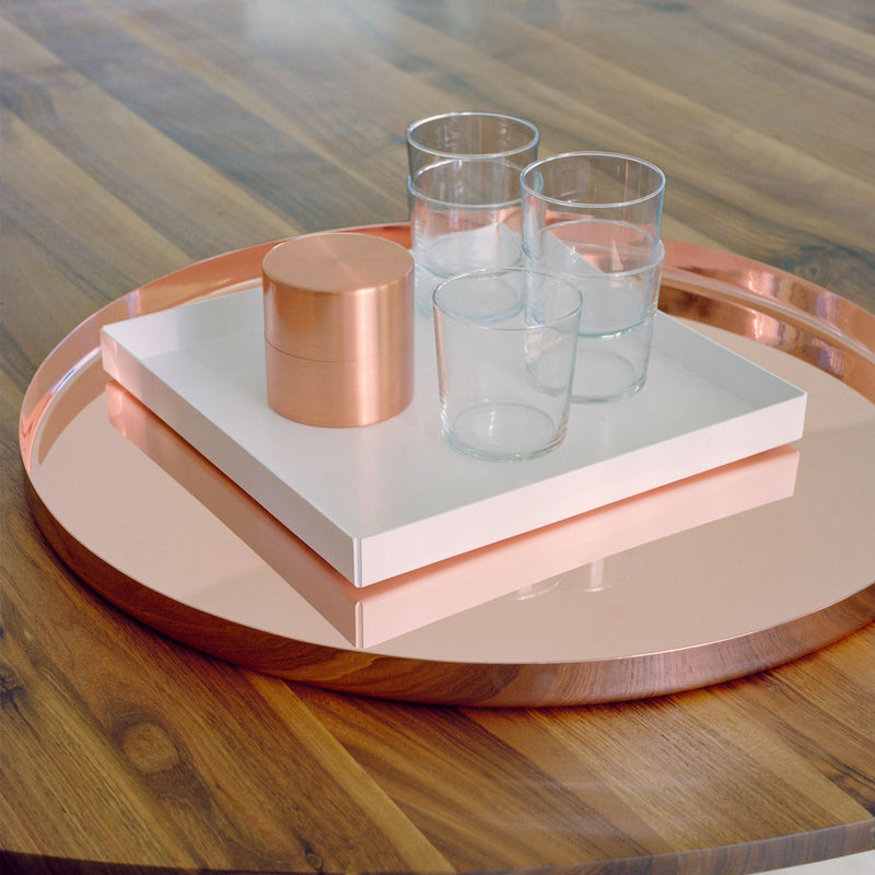 A round wooden side table features the E15 CM05 HABIBI TRAY with a rose gold finish, holding three clear glass tumblers and a rose gold container, all beautifully arranged on a white rectangular board.