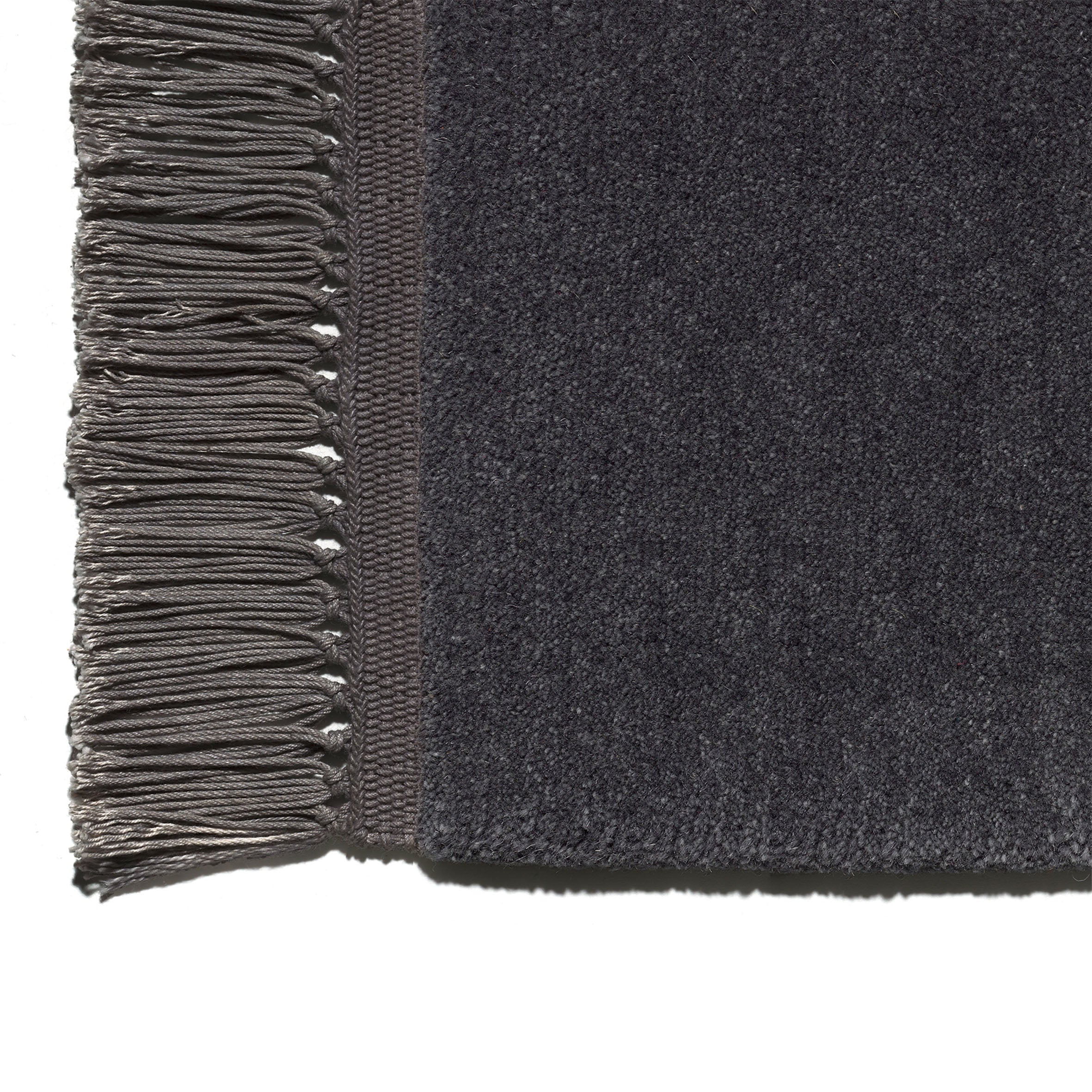 A close-up of the CP03 KAVIR by E15 showcases a dark gray, textured, hand-woven carpet with a decorative fringe on the left made of tightly twisted threads. Its main body features a soft, plush surface.
