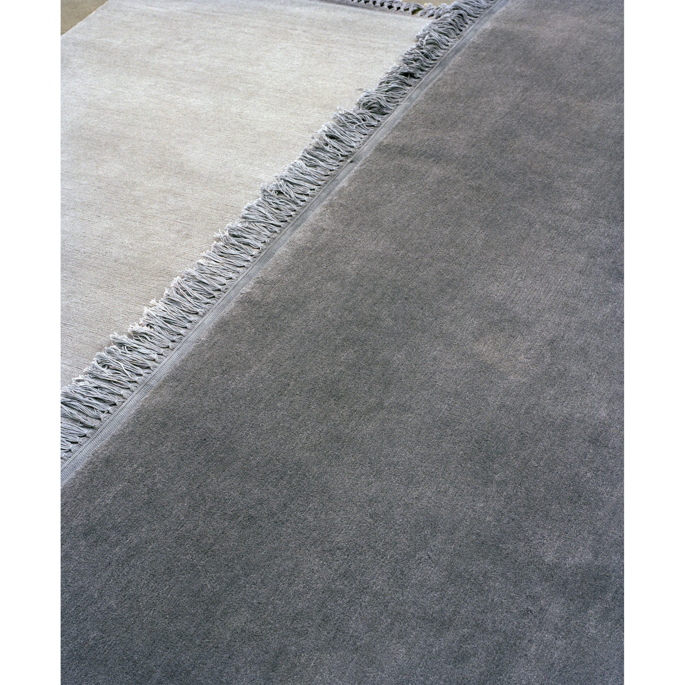 Close-up of overlapping carpets on a floor. The E15 CP03 KAVIR, a hand-woven Persian carpet, appears in light gray with fringed edges on the left, and a dark gray one is seen on the right. Textures and subtle color differences are highlighted in the image.
