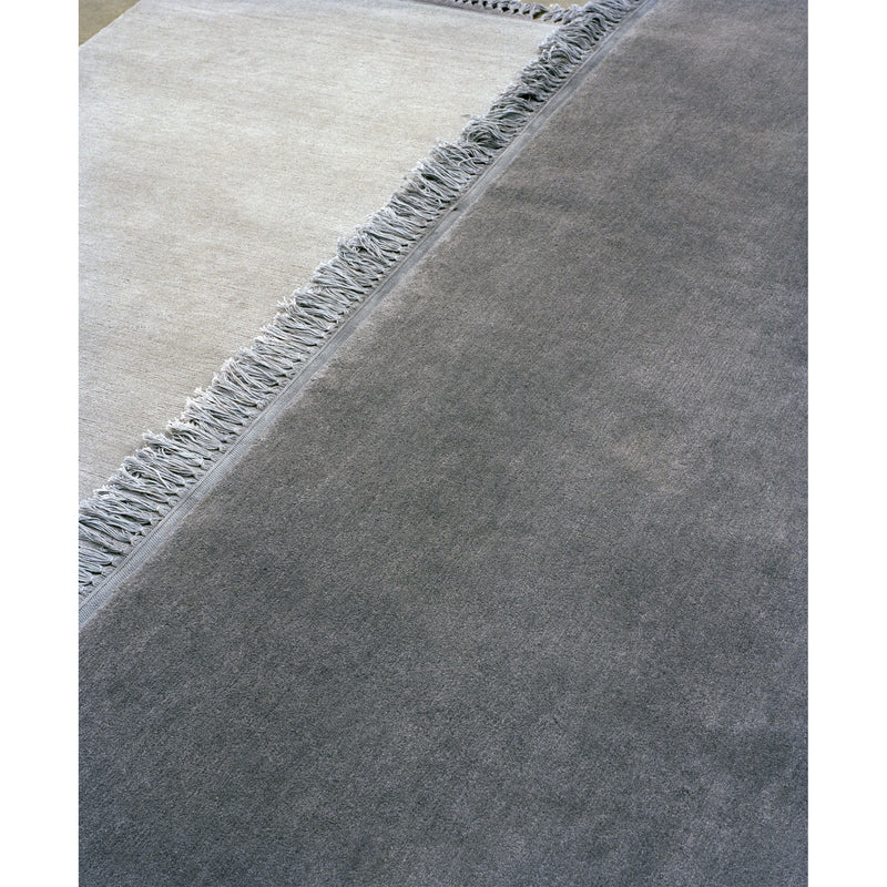 Close-up of overlapping carpets on a floor. The E15 CP03 KAVIR, a hand-woven Persian carpet, appears in light gray with fringed edges on the left, and a dark gray one is seen on the right. Textures and subtle color differences are highlighted in the image.