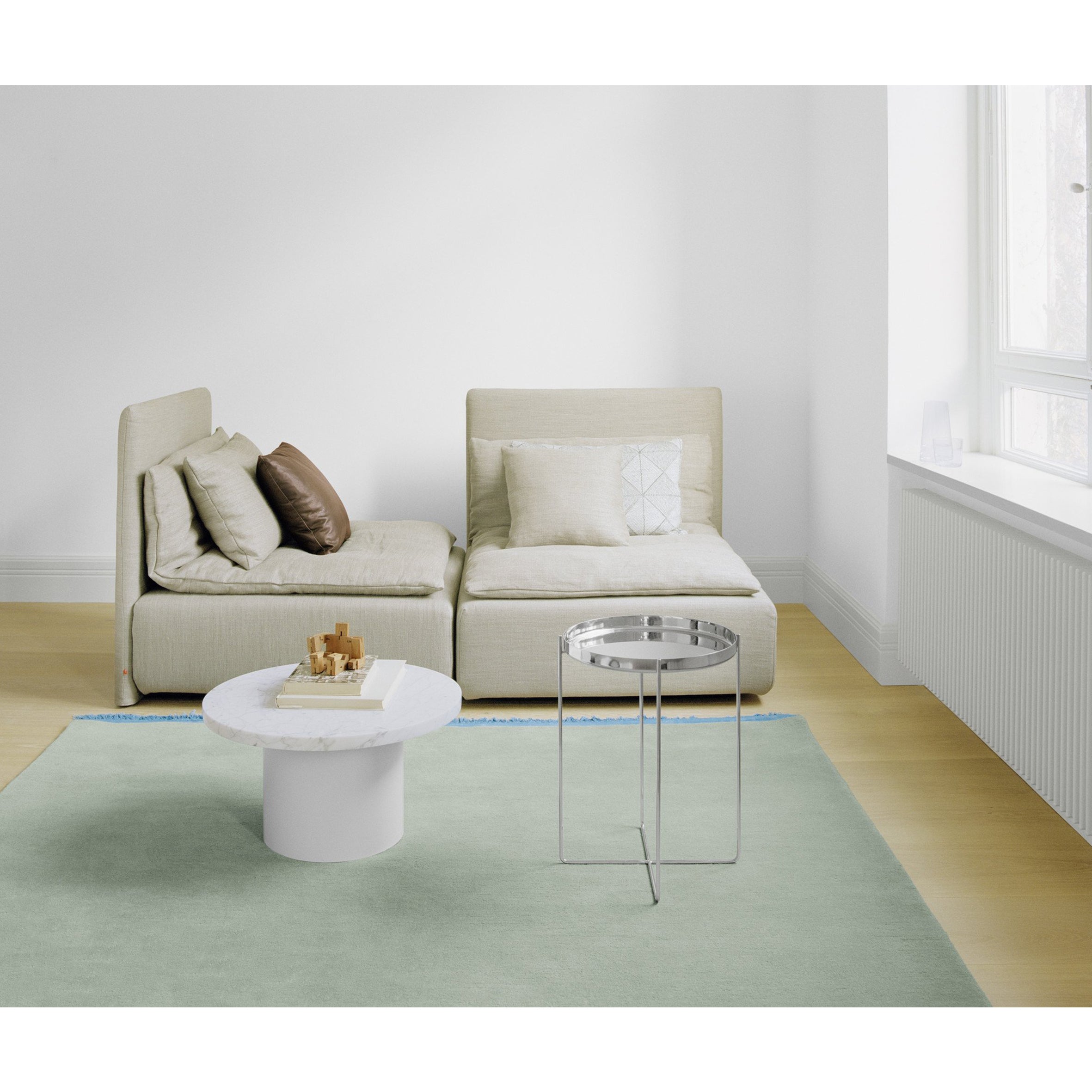 A minimalist living room features a beige modular sofa, light blue and beige cushions, a white marble coffee table, and a sleek round metal side table. The hand-woven CP03 KAVIR carpet by E15 enhances the space, while large windows flood it with natural light.