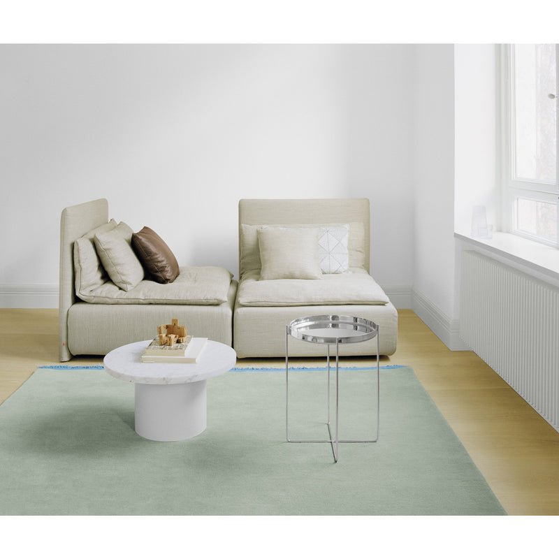 A minimalist living room features a beige modular sofa, light blue and beige cushions, a white marble coffee table, and a sleek round metal side table. The hand-woven CP03 KAVIR carpet by E15 enhances the space, while large windows flood it with natural light.