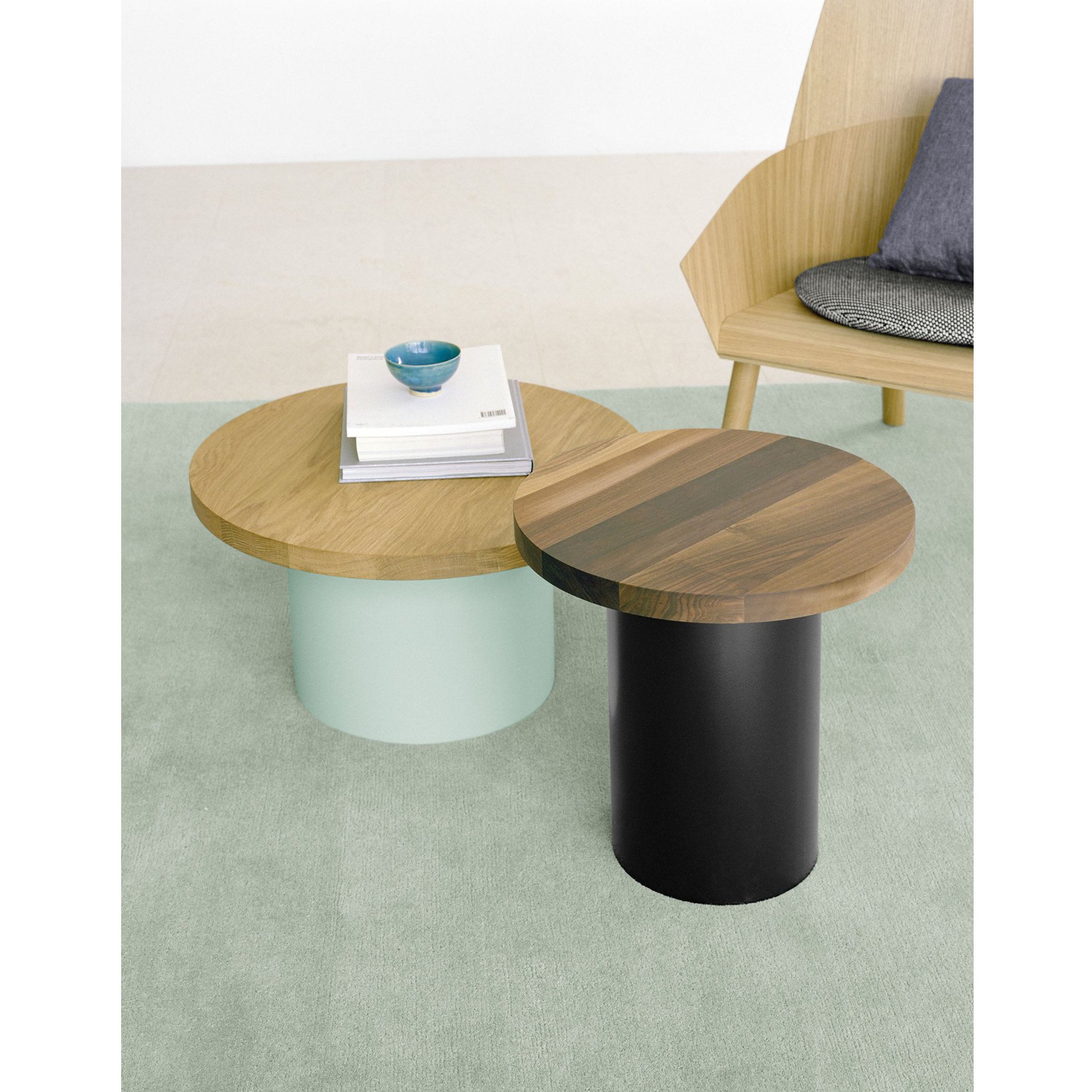 The CP03 KAVIR by E15, featuring two round wooden nesting tables with cylindrical bases (mint green and black), rests on a hand-woven Persian carpet. A modern wooden chair with a gray cushion accompanies a stack of books and a blue bowl elegantly placed on the larger table.