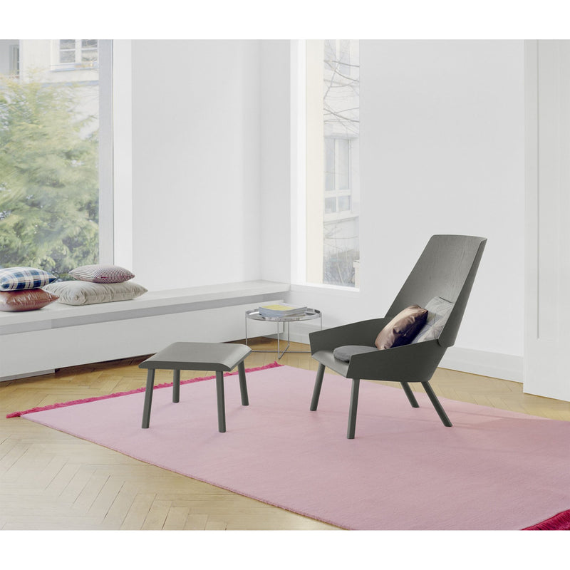 A modern minimalist room includes a tall gray chair with matching footstool on an E15 CP03 KAVIR hand-woven carpet. A round side table is nearby, and a bench with colorful pillows rests by a large window showcasing lush greenery.