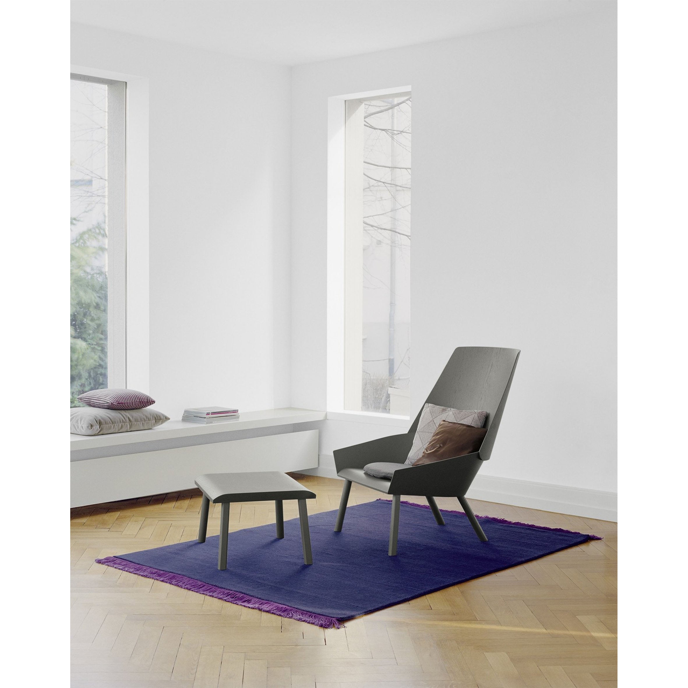 A modern living room showcases the CP03 KAVIR by E15, with a gray chair and matching footstool on a hand-woven carpet. Large windows fill the light wood parquet floor space with natural brightness, while cushions adorn a built-in bench by the window.
