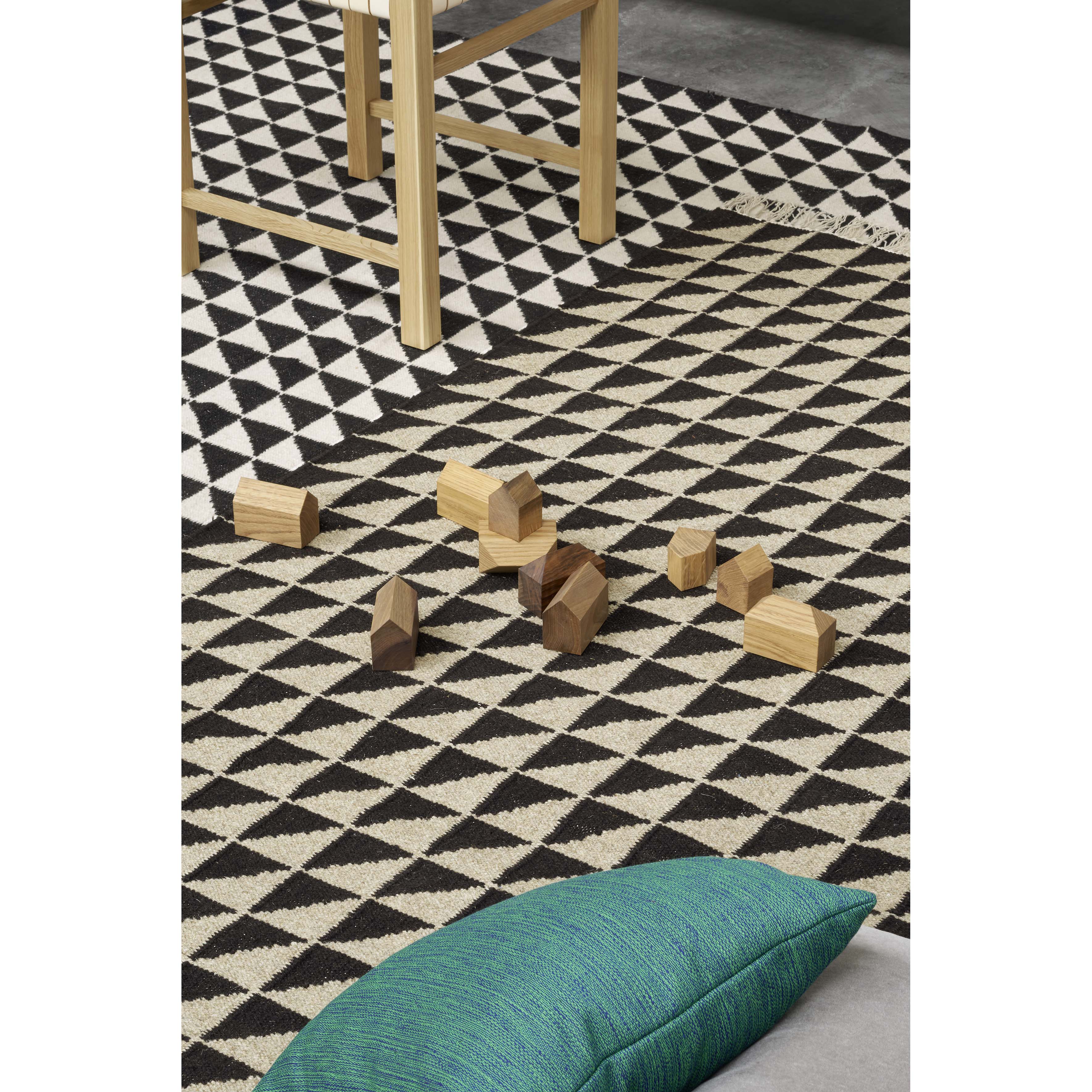 The CP04 IZA from E15 is a patterned black and white rug made of hand-spun lambs wool with scattered wooden blocks. A wooden chair is partially visible, complemented by a green cushion in the foreground, highlighting E15s focus on detail and traditional weaving.