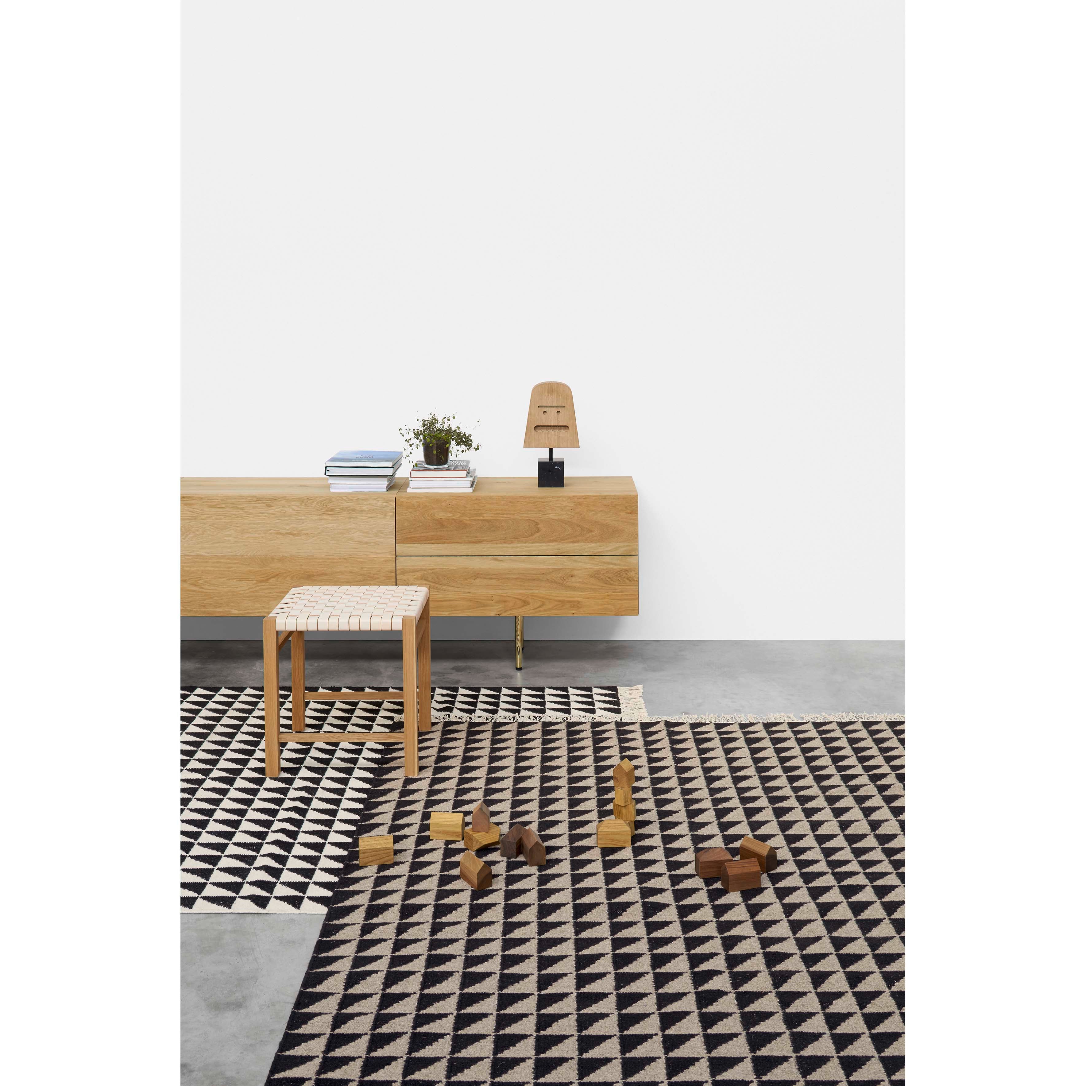 A modern minimalist room features a CP04 IZA wooden sideboard and stool by E15 on a traditional black and white geometric rug, topped with a table lamp and books. Wooden blocks scattered on the floor add texture to the designer-inspired space.