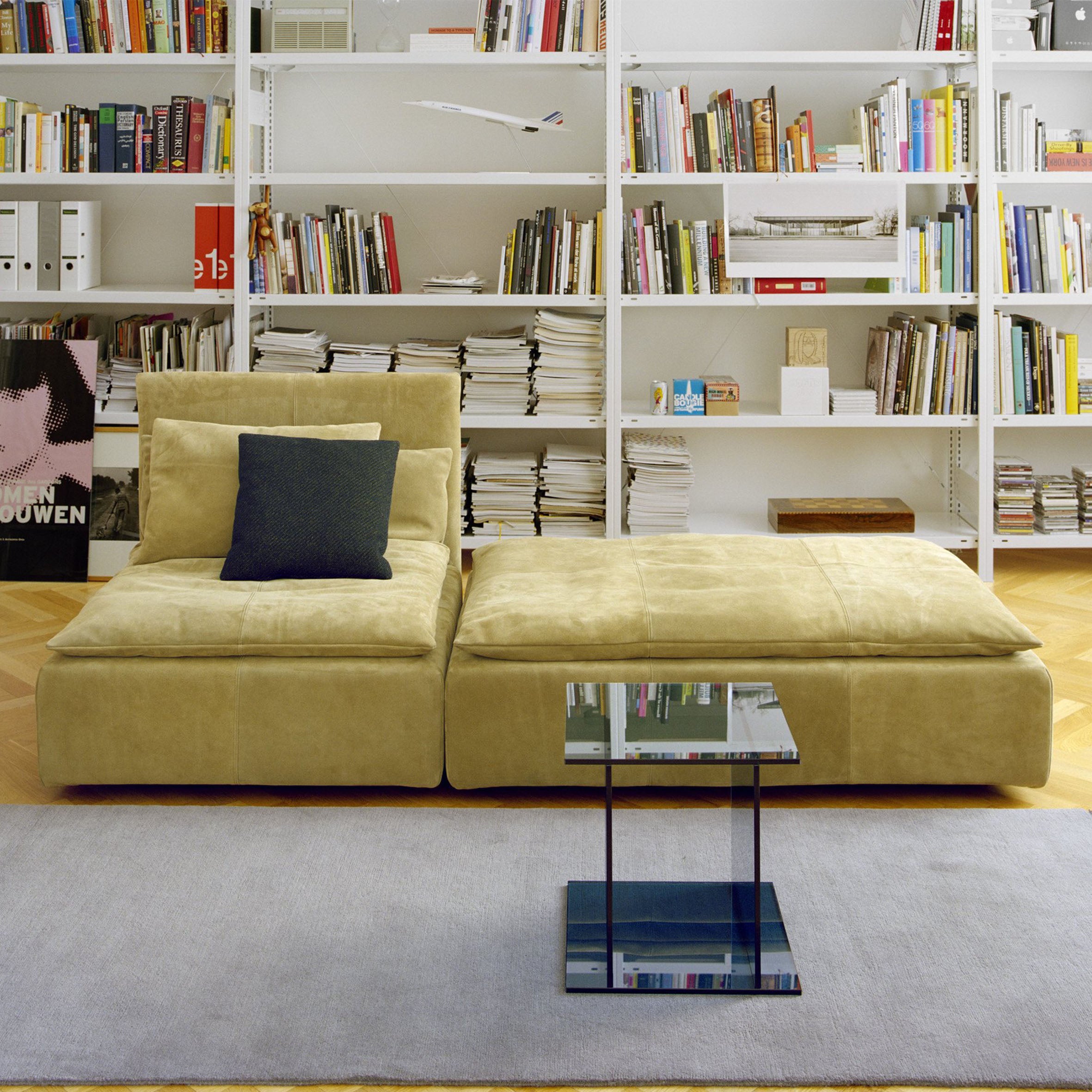 In a modern living room, the mustard yellow sectional sofa features a dark cushion. The CT07 Drei glass side table by E15 stands in front, backed by bookshelves filled with storage. A cozy gray area rug complements the hardwood floor.