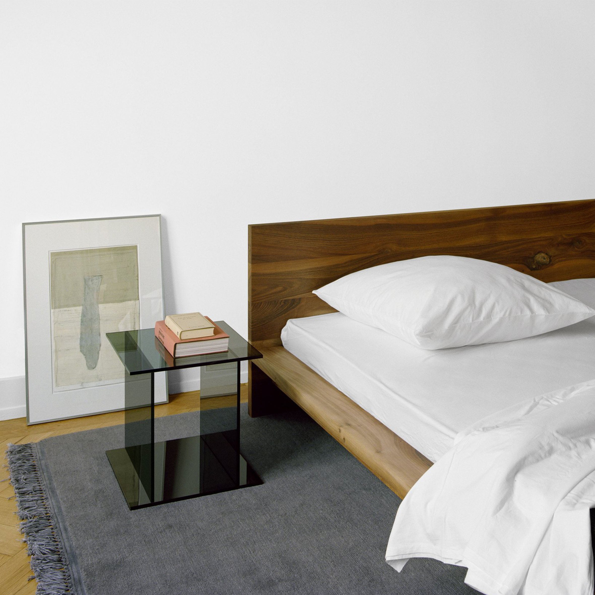 The minimalist bedroom showcases a wooden CT07 Drei bed by E15 with white bedding and a gray rug. A sleek black glass side table provides ample book storage. A framed abstract art piece leans against the white wall, while the light wood floor enhances the modern, serene atmosphere.