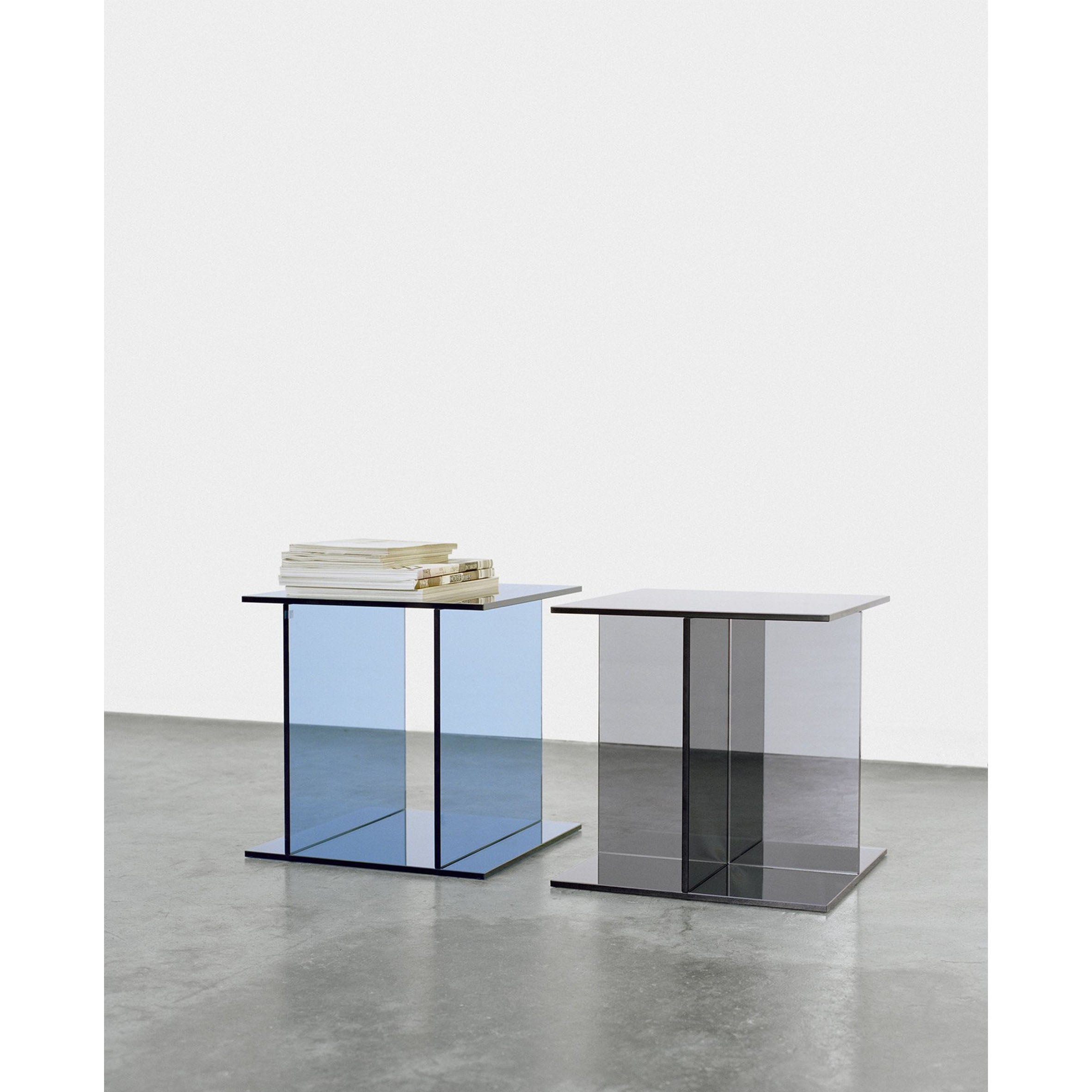 Two modern, transparent CT08 VIER tables by E15 with square tops rest on a gray concrete floor. One is crafted from solid colored glass and holds a small stack of books, with subtle hues reflecting from their surfaces and one showing a blue tint.