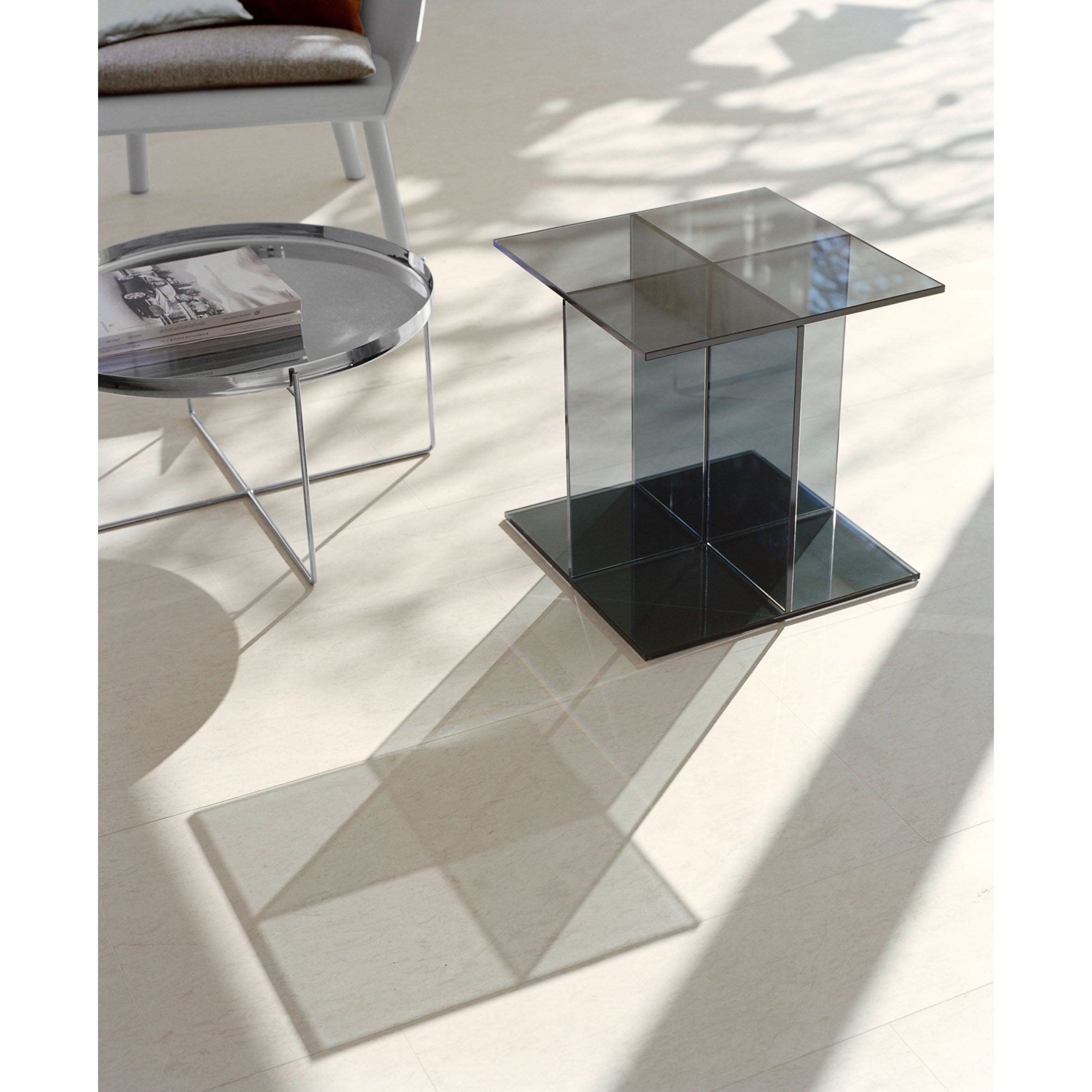 The E15 CT08 VIER modern glass table casts a shadow on a light floor in a sunlit room. Nearby, a round metal tray holds papers, and a chair with a cushion peeks out in the background, highlighting the sleek design of the table.